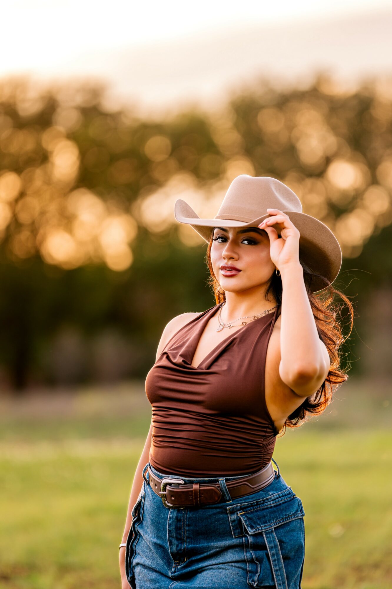 Woman wearing a wide-brimmed hat and brown top standing outdoors with trees and sunset in background.