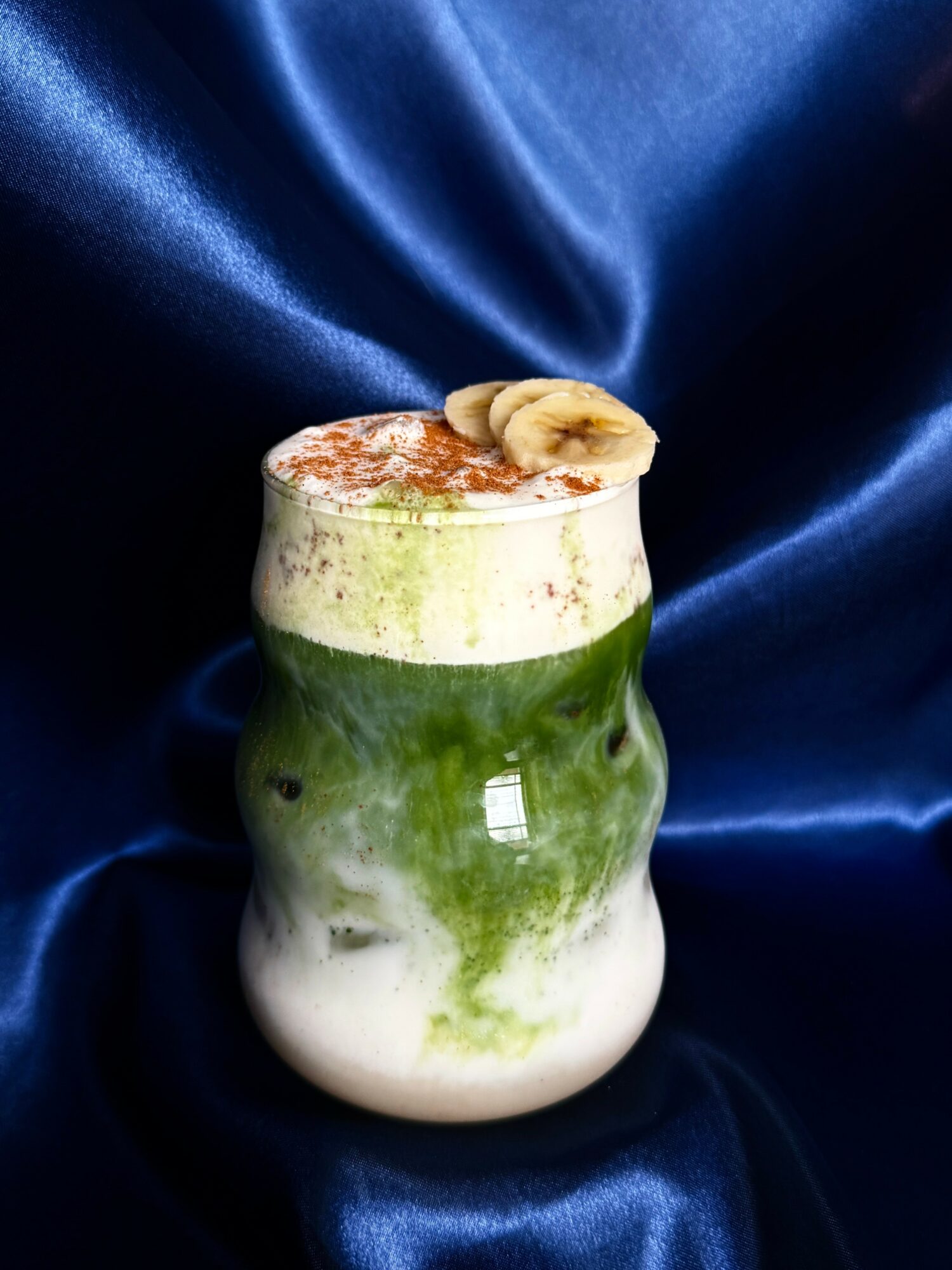 A layered green and white smoothie in a glass with banana slices on top, set against a blue satin background.