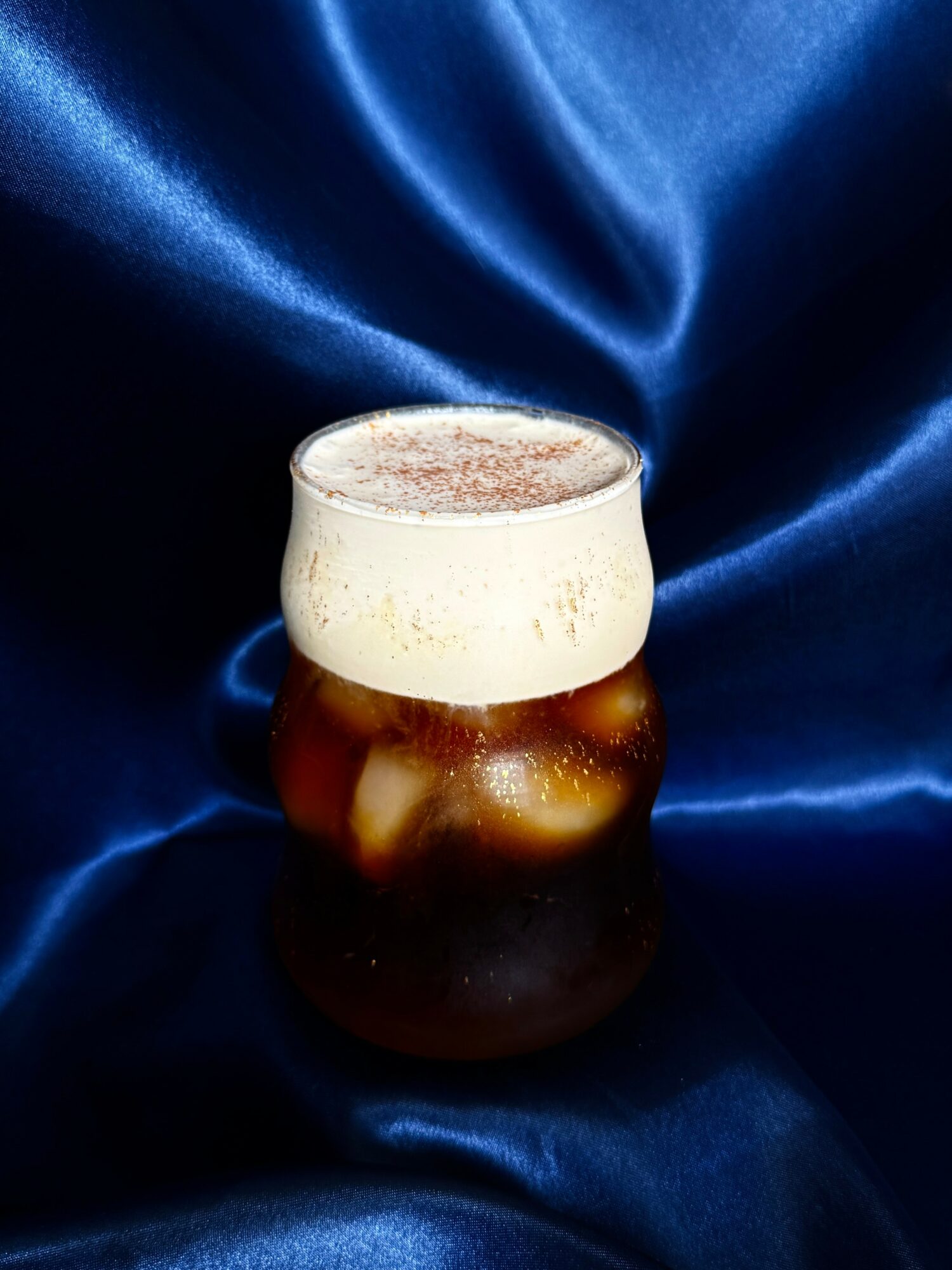 A glass of dark beer with a frothy head on a shiny blue fabric background.