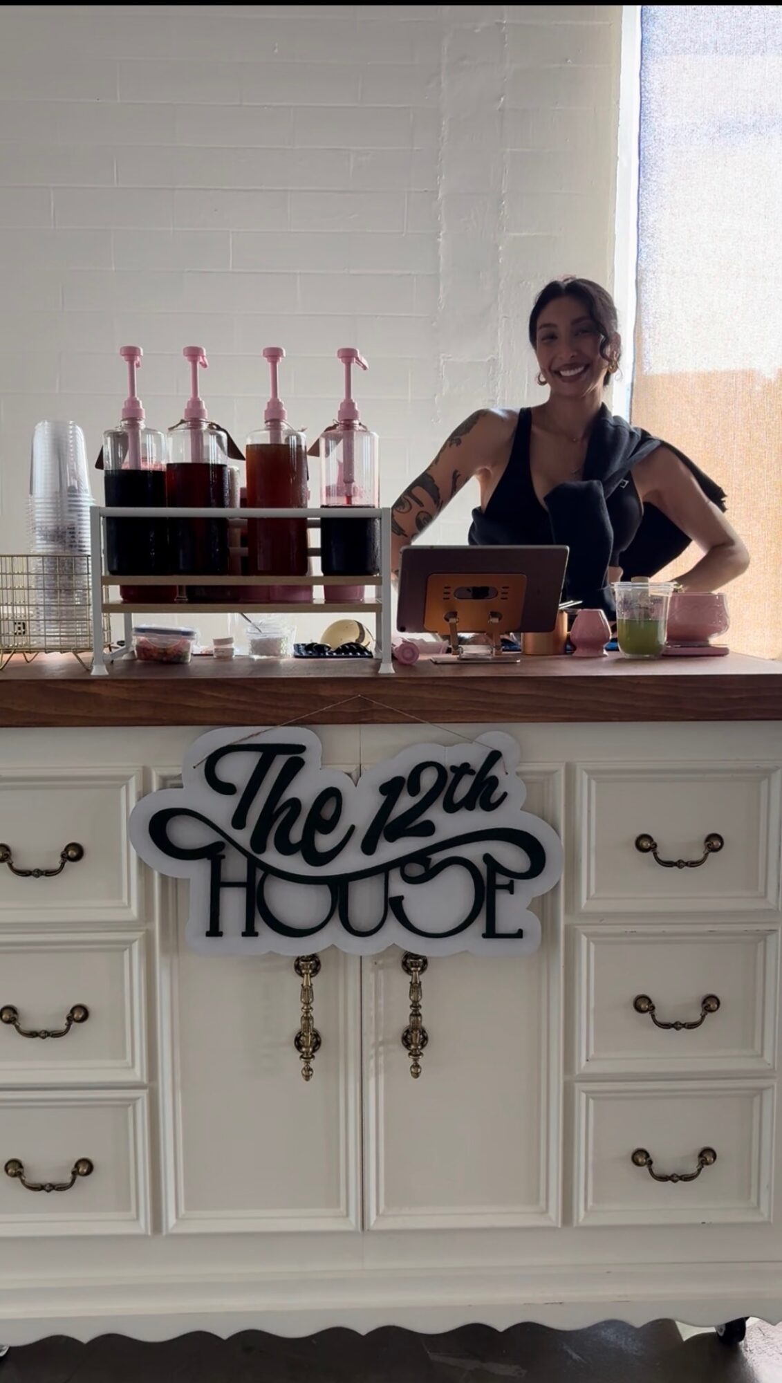 A woman stands behind a counter with a sign reading 'The 12th House,' in a bright room with a large window.
