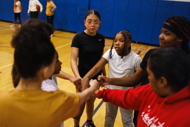 Group of children holding hands in a gymnasium, some facing the camera and others with backs turned.