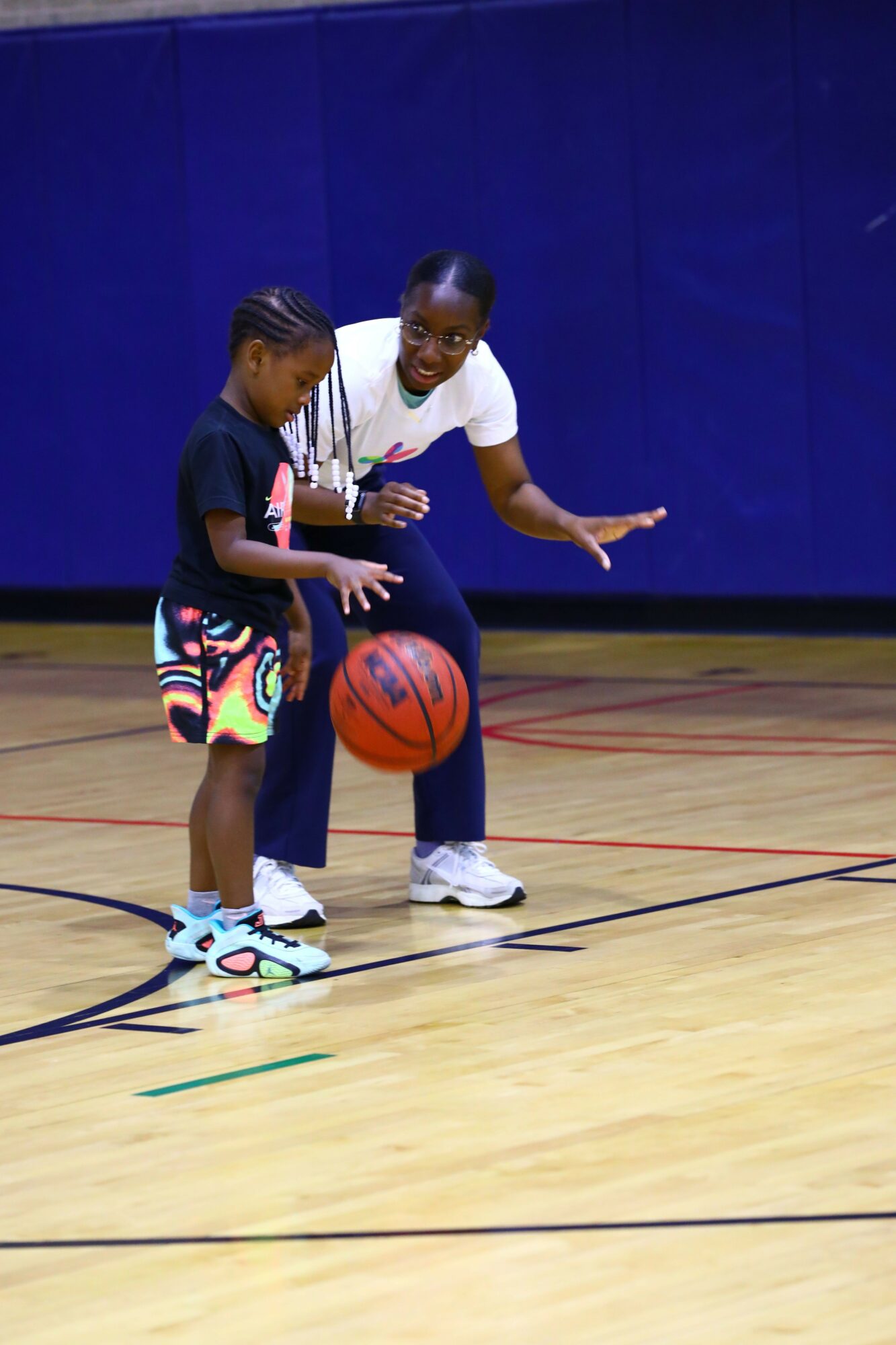 Two women on a basketball court, one holding a basketball, with a blue wall in the background.