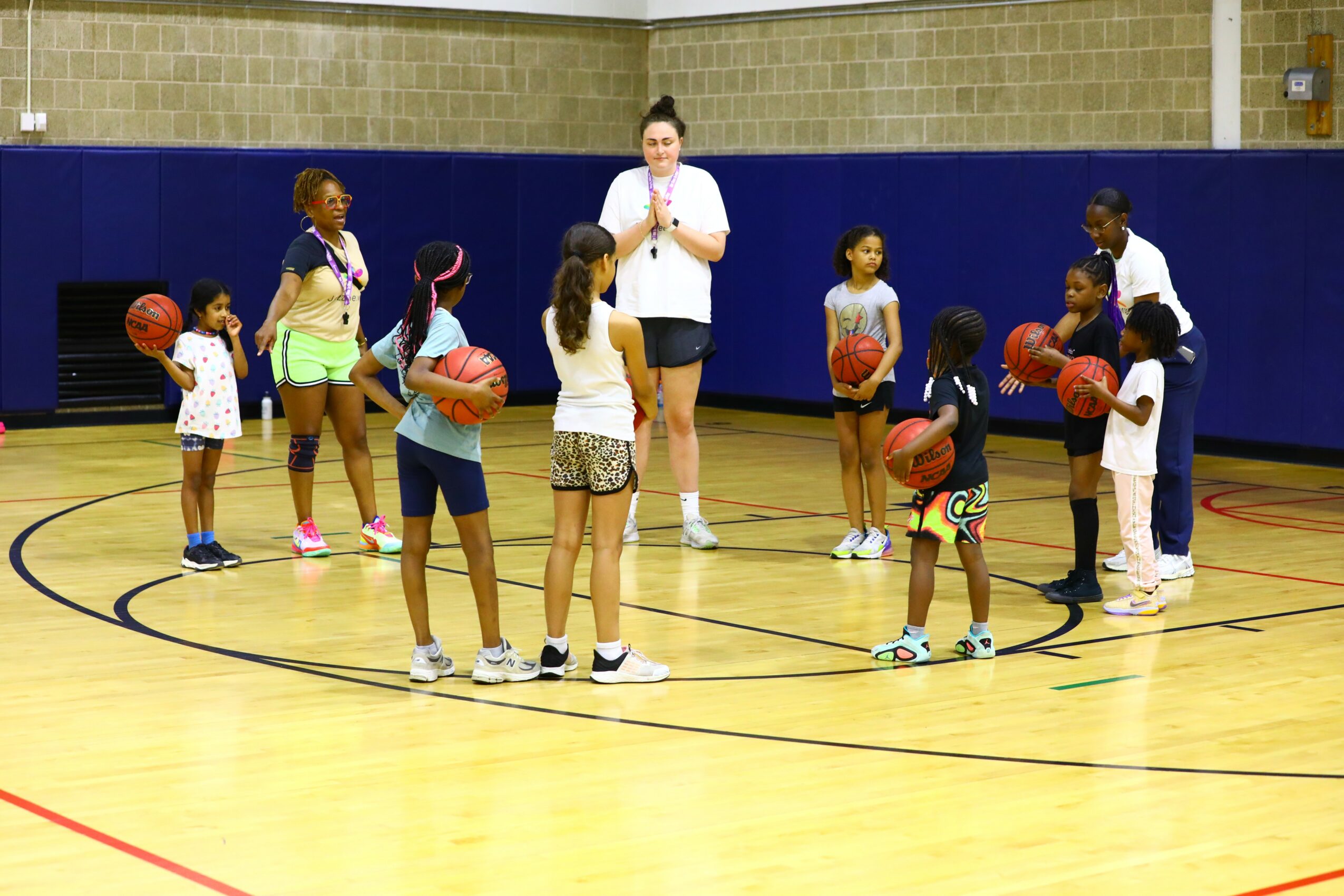 Group of children and two adults in a gymnasium, holding basketballs, listening to a coach, with a blue wall background.