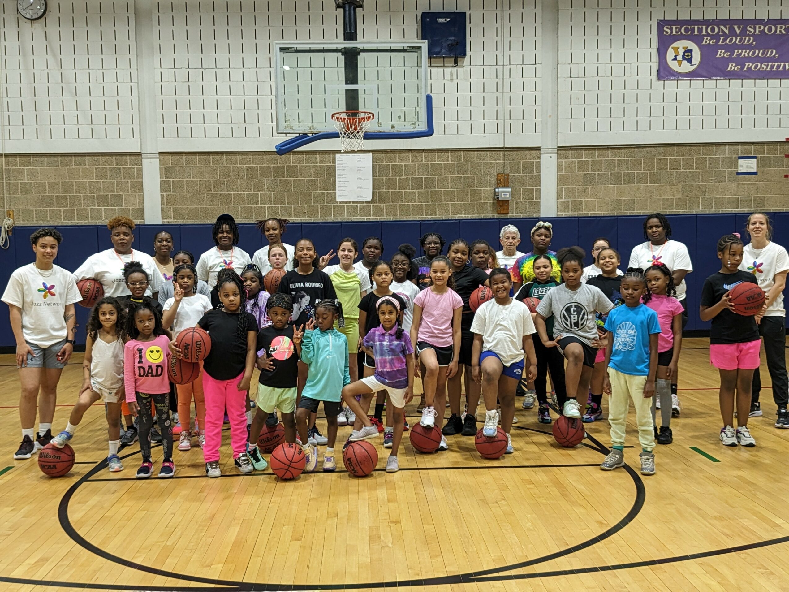 Group of children and adults in a gymnasium, some holding basketballs, standing on a basketball court with a hoop above.