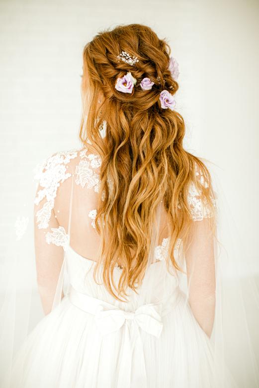 Back view of a woman with long, wavy red hair decorated with flowers, wearing a white lace dress.