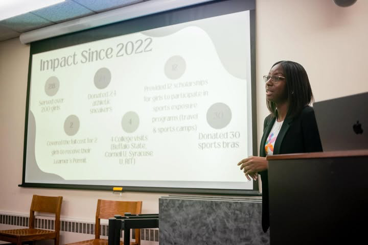 A woman stands in front of a presentation screen displaying text and graphics in a classroom or conference room.
