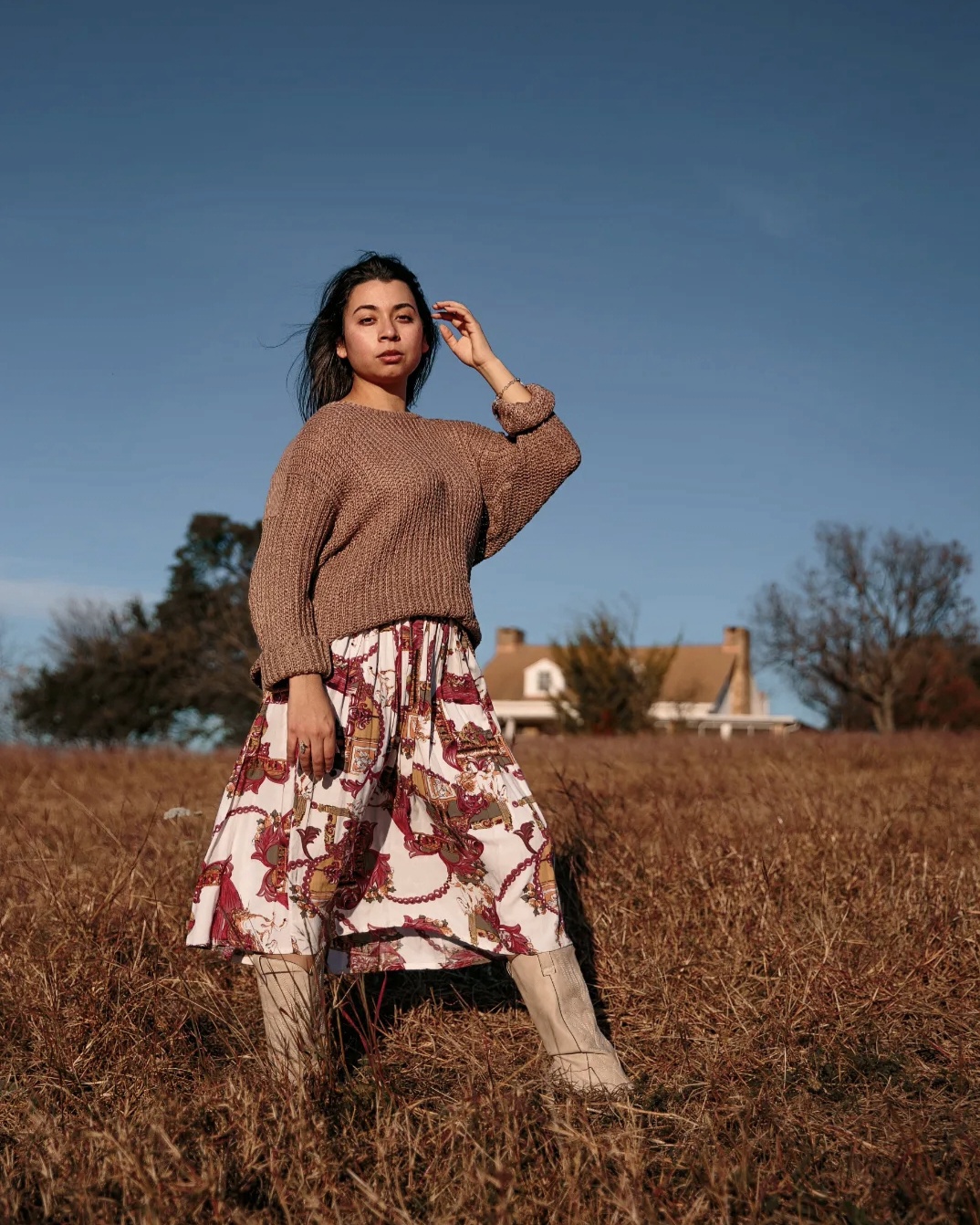 Young woman standing in a field under a clear blue sky, wearing a brown sweater and patterned skirt, with trees and houses in background.