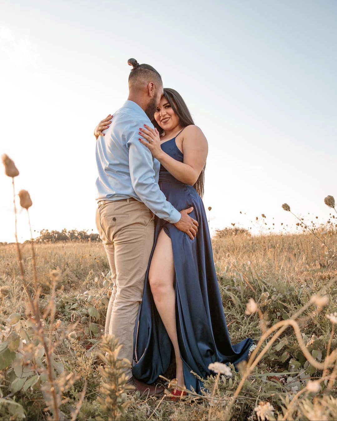 A couple hugging in a field, woman in a blue dress, man in a light shirt and beige pants, clear sky background.