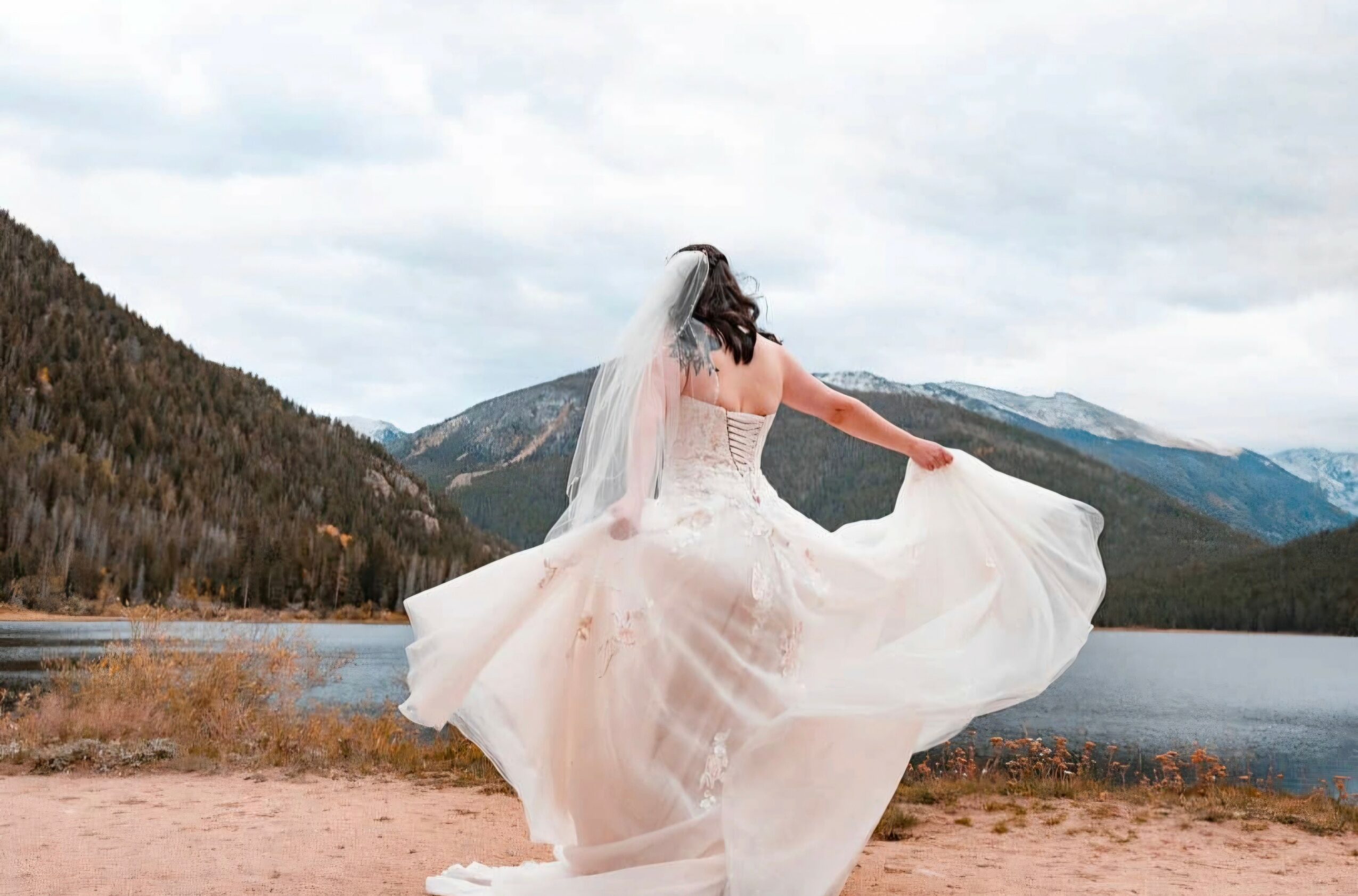 Woman in wedding dress and veil outdoors near a lake with mountains in the background, holding her dress out.