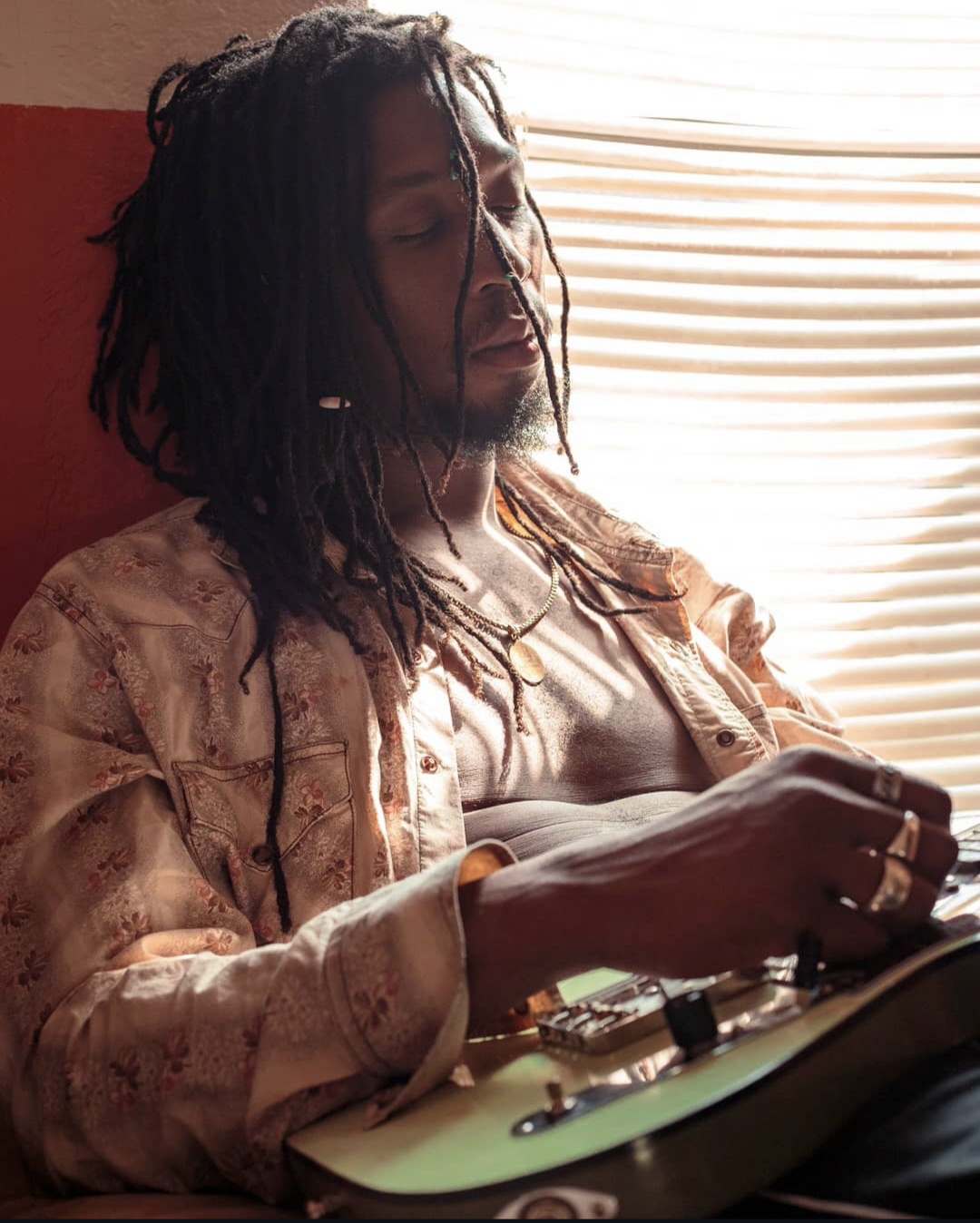 Person with dreadlocks sitting with eyes closed, wearing a patterned shirt, near window with blinds, holding a phone.