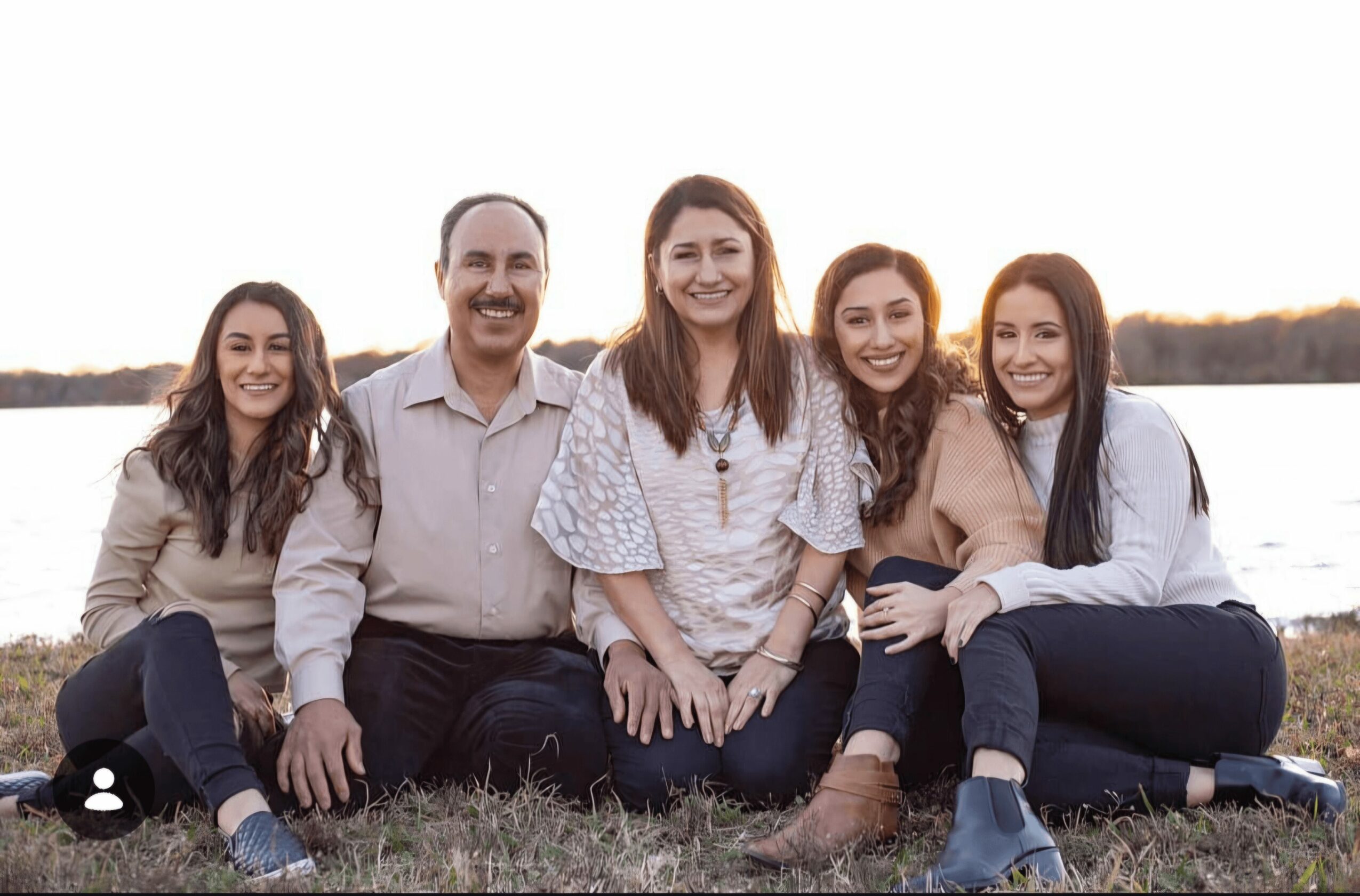 Group of five people sitting outdoors near water, smiling, with a sunset in the background.