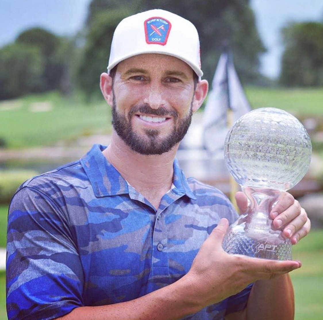 Man with a beard holding a glass trophy outdoors, smiling, wearing a white cap and blue patterned shirt.