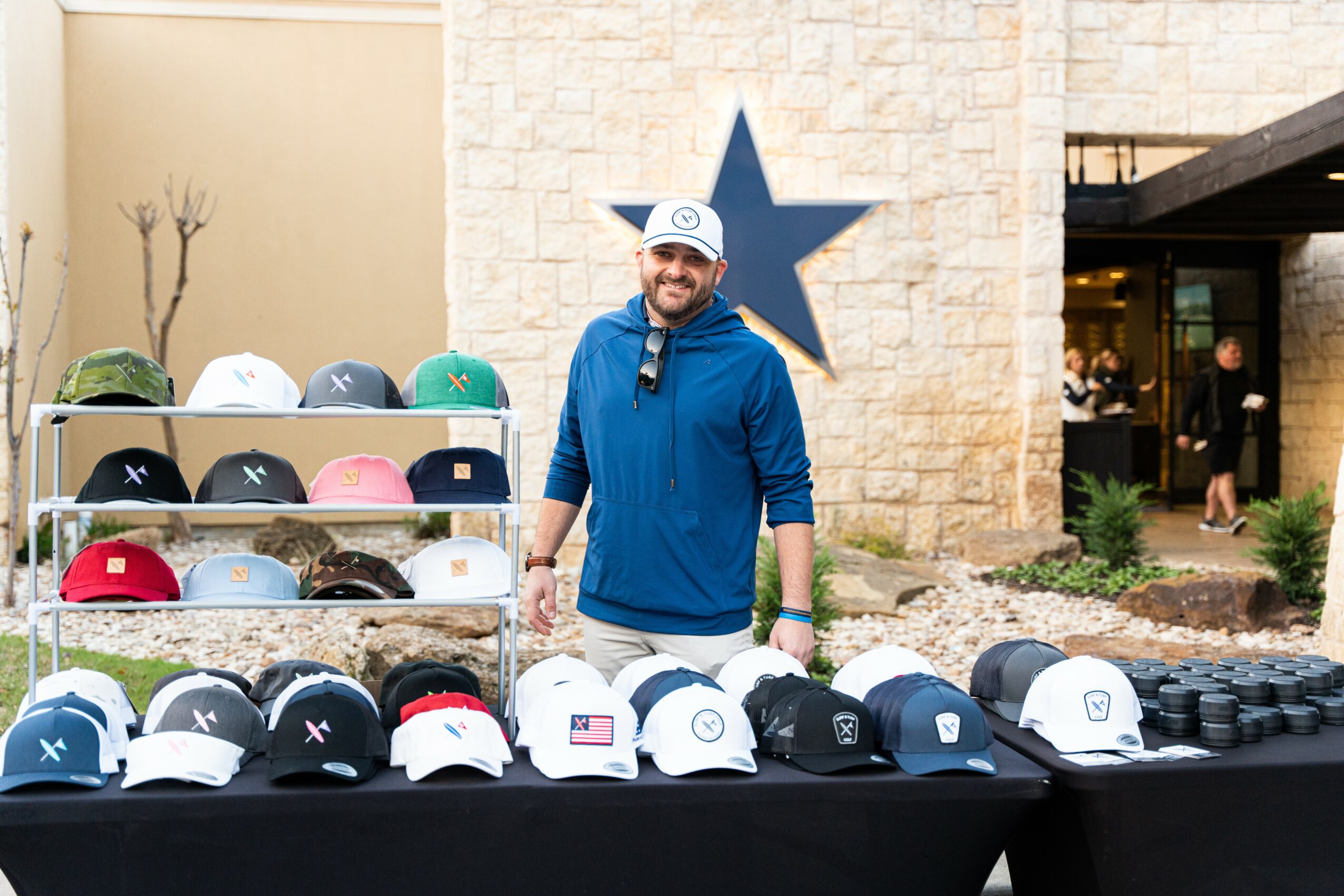 Man in blue hoodie standing behind a table of hats outdoors, with a large star on a wall behind him.
