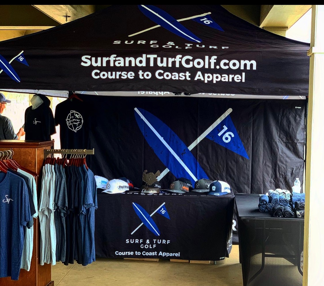 Golf merchandise booth with hats and shirts, branded SurfundTurfGolf.com, under a black canopy with blue and white graphics.