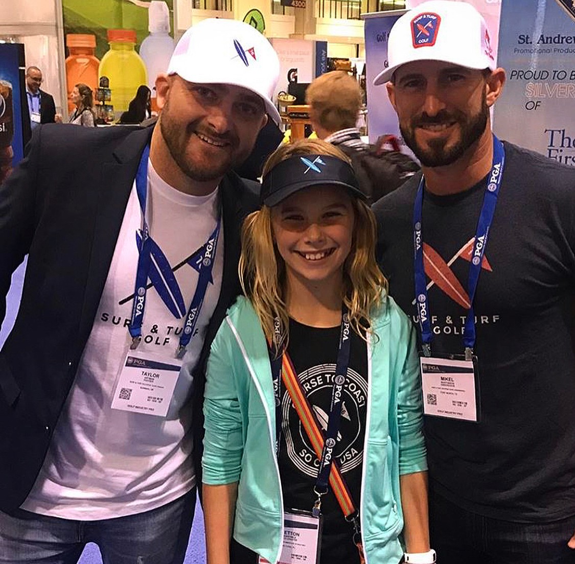 Two men and a girl smiling at an event, wearing caps and badges, with colorful background and banners.