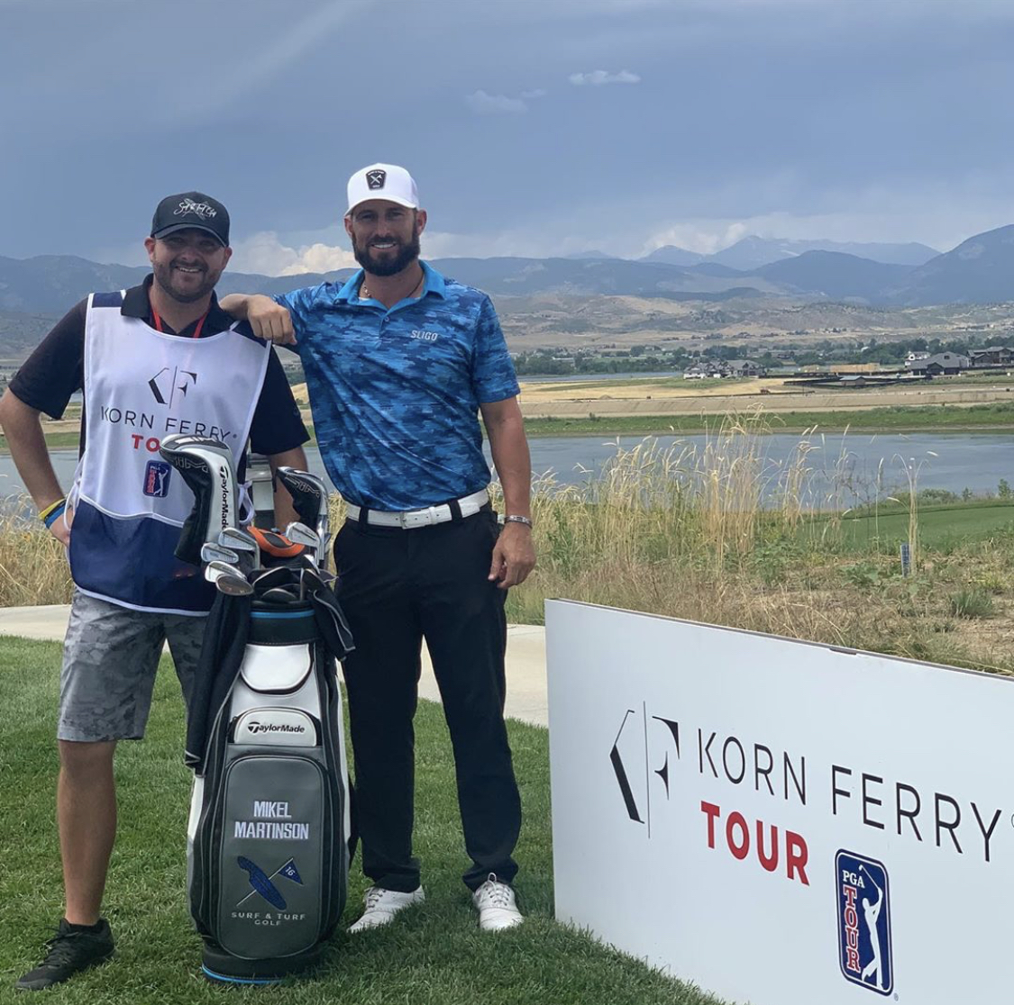 Two men standing outdoors near a golf course with a sign that reads 'Korn Ferry Tour'. One man has a golf bag, and they are smiling.