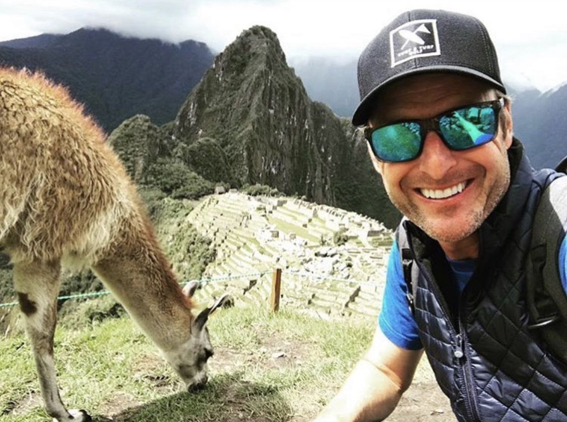 Smiling man wearing sunglasses and a cap, with a llama and Machu Picchu in the background.