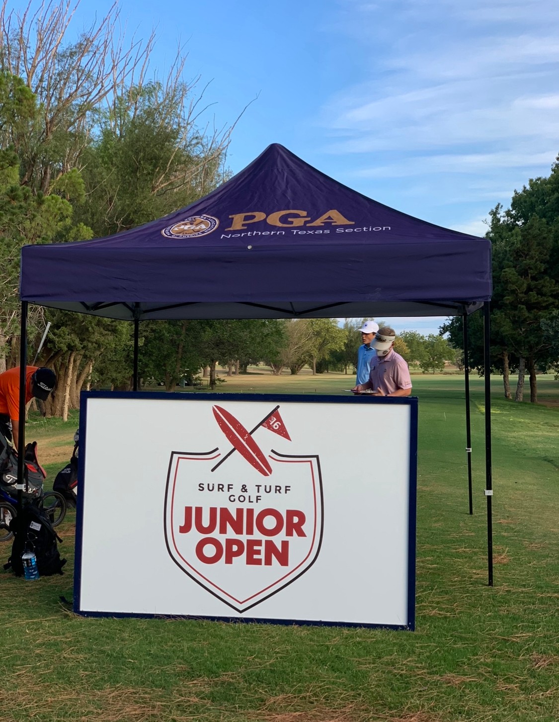 A blue PGA tent with a white sign reading 'Junior Open' on a golf course, with people and trees in the background.