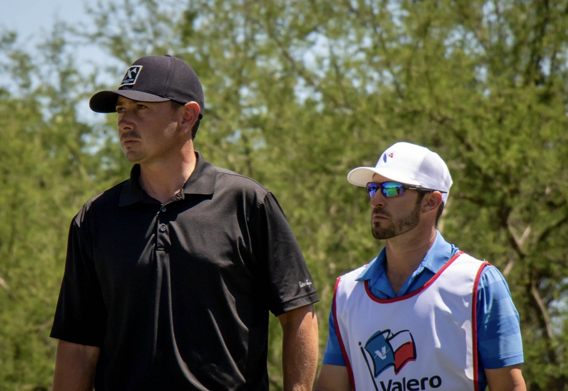 Two men outdoors, one in a black shirt and cap, the other in a white cap and vest, with trees in background.