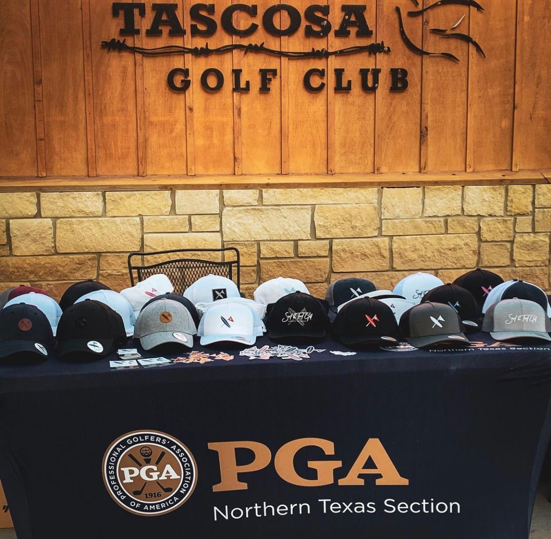 Table with golf hats displayed in front of a wooden wall with a sign for Tascosa Golf Club, PGA Northern Texas Section logo, and hats in various colors.