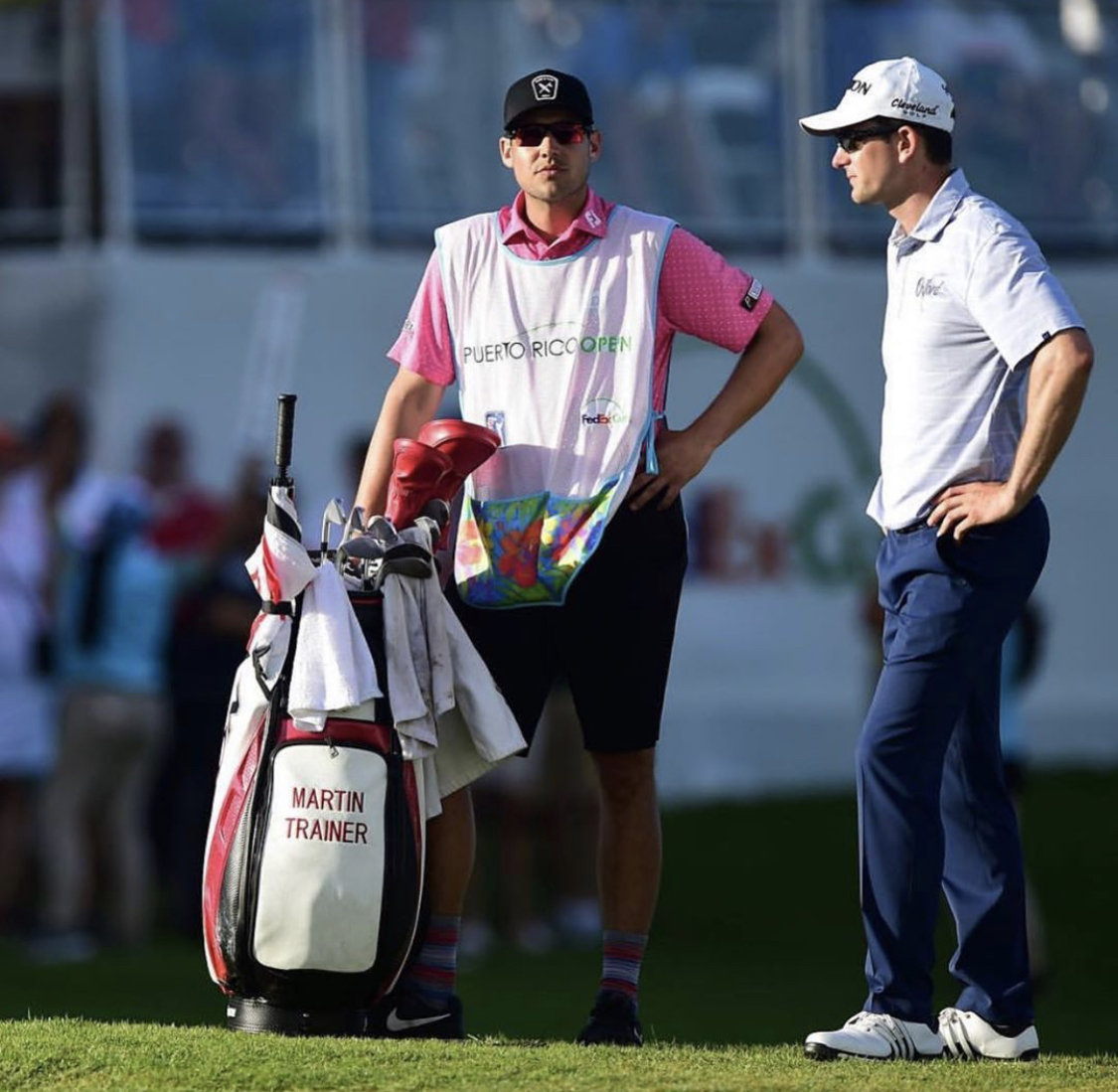 Two men standing on a golf course, one with a golf bag labeled 'Martin Trainer,' talking outdoors.