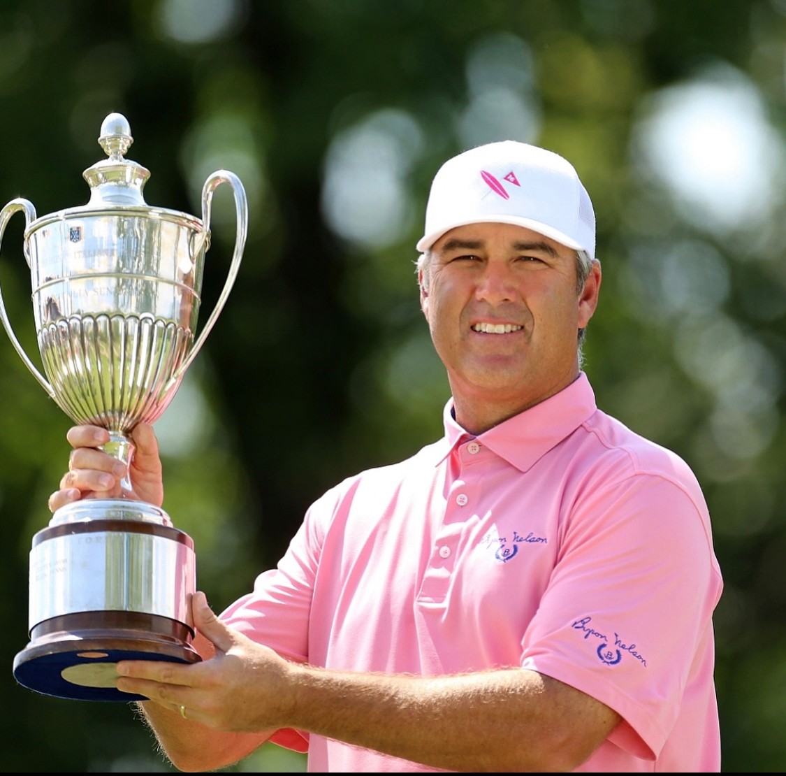Man in pink polo shirt and white cap holding a large silver trophy outdoors, smiling, with blurred green trees in background.