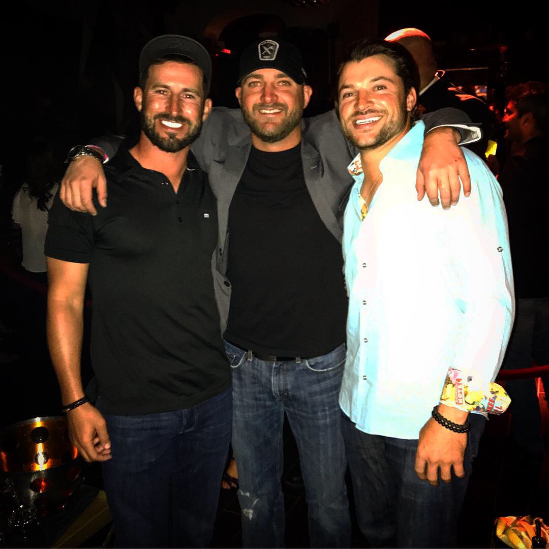 Three men smiling and posing together in a dark indoor setting, with arms around each other, wearing casual clothing.