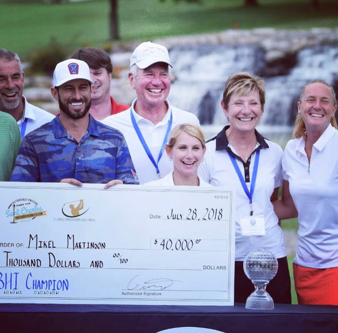 Group of smiling people holding a large check outdoors near a waterfall, celebrating a victory or award, with a trophy in front.