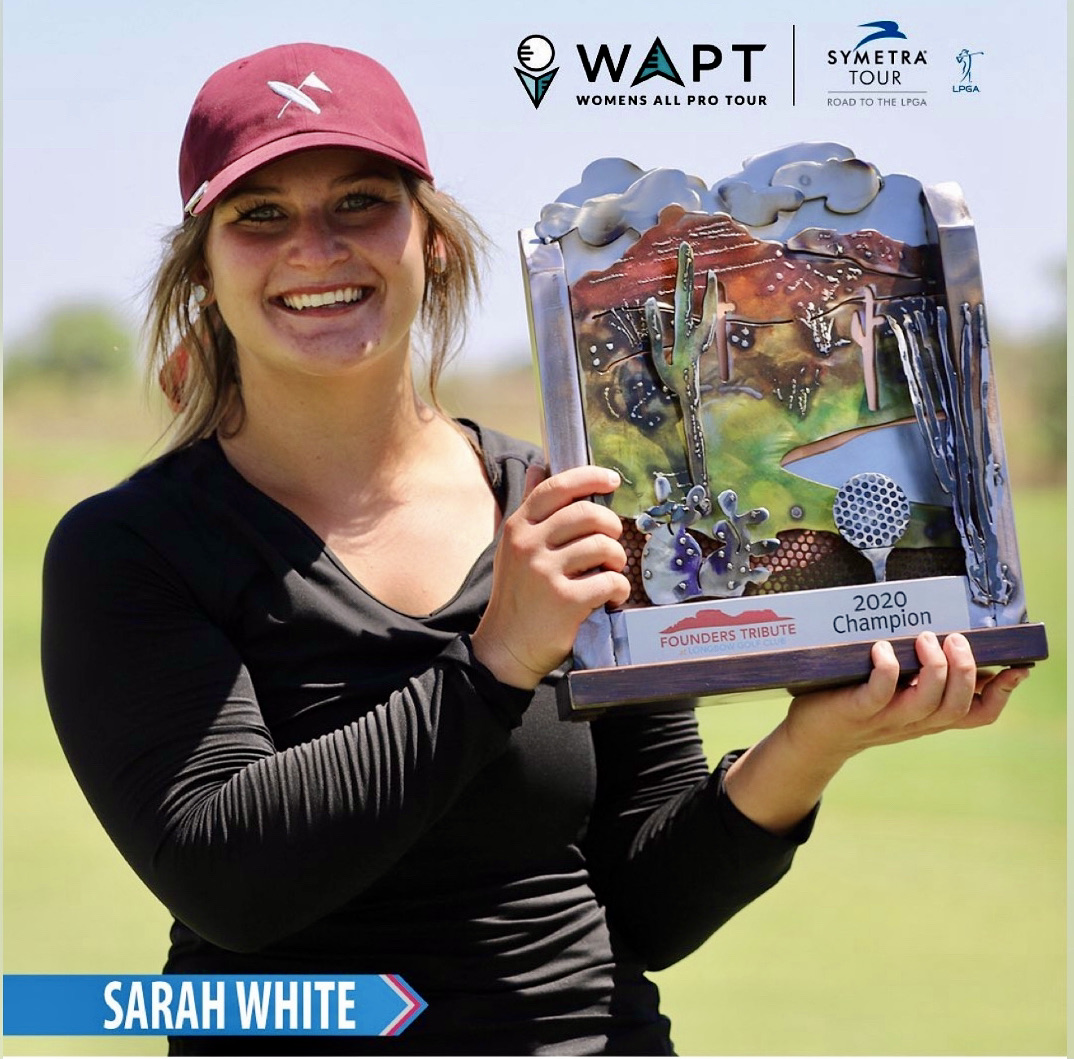 Woman smiling holding a trophy with a golf course background, wearing a pink cap and black shirt.