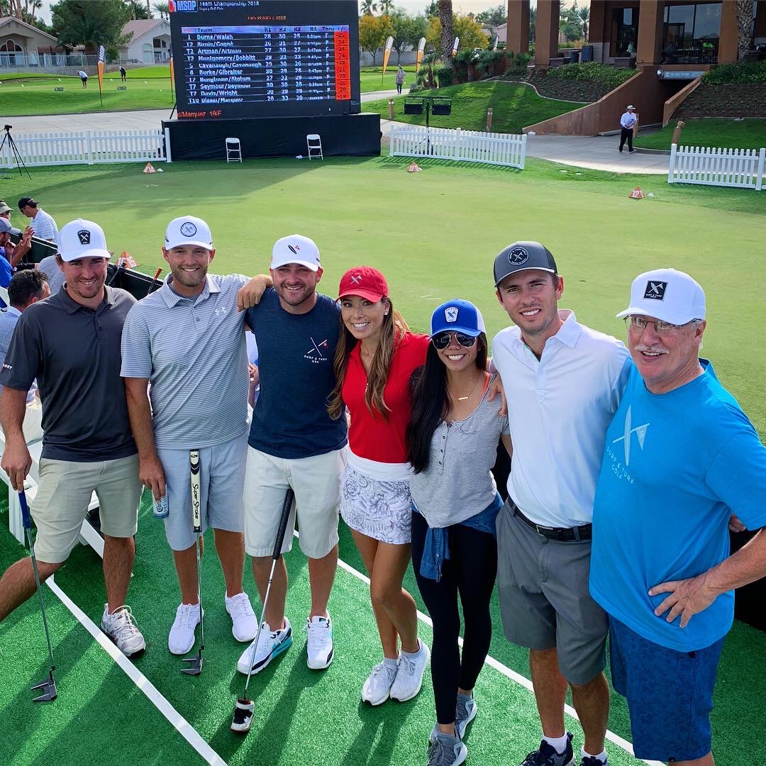 Group of seven people smiling on a golf course, standing in front of a scoreboard and white fence, sunny day.