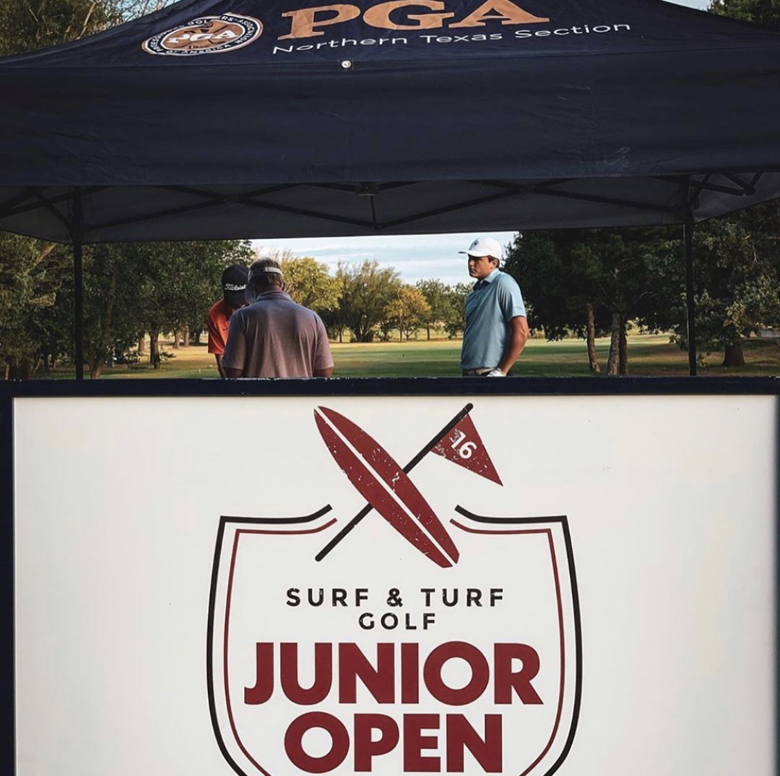 People under a tent at a golf event, with a sign reading 'Junior Open' and a surfboard logo.