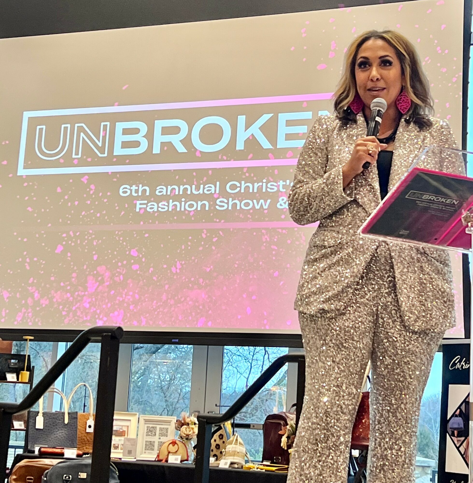 Woman speaking into microphone at event, holding a pink folder, with a large screen behind displaying event details.