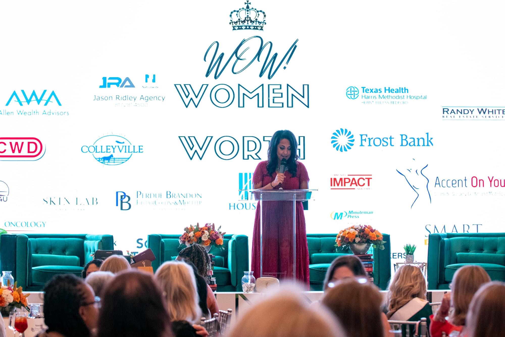 Woman speaking at a podium during a women’s event, with a large screen behind displaying logos and text, audience seated.
