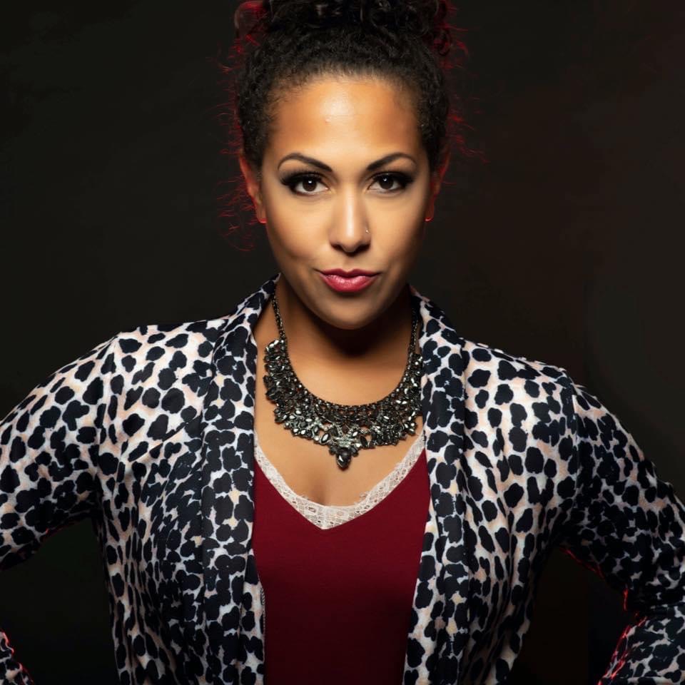 Woman with dark curly hair, wearing a leopard print jacket and a statement necklace, poses confidently against a dark background.