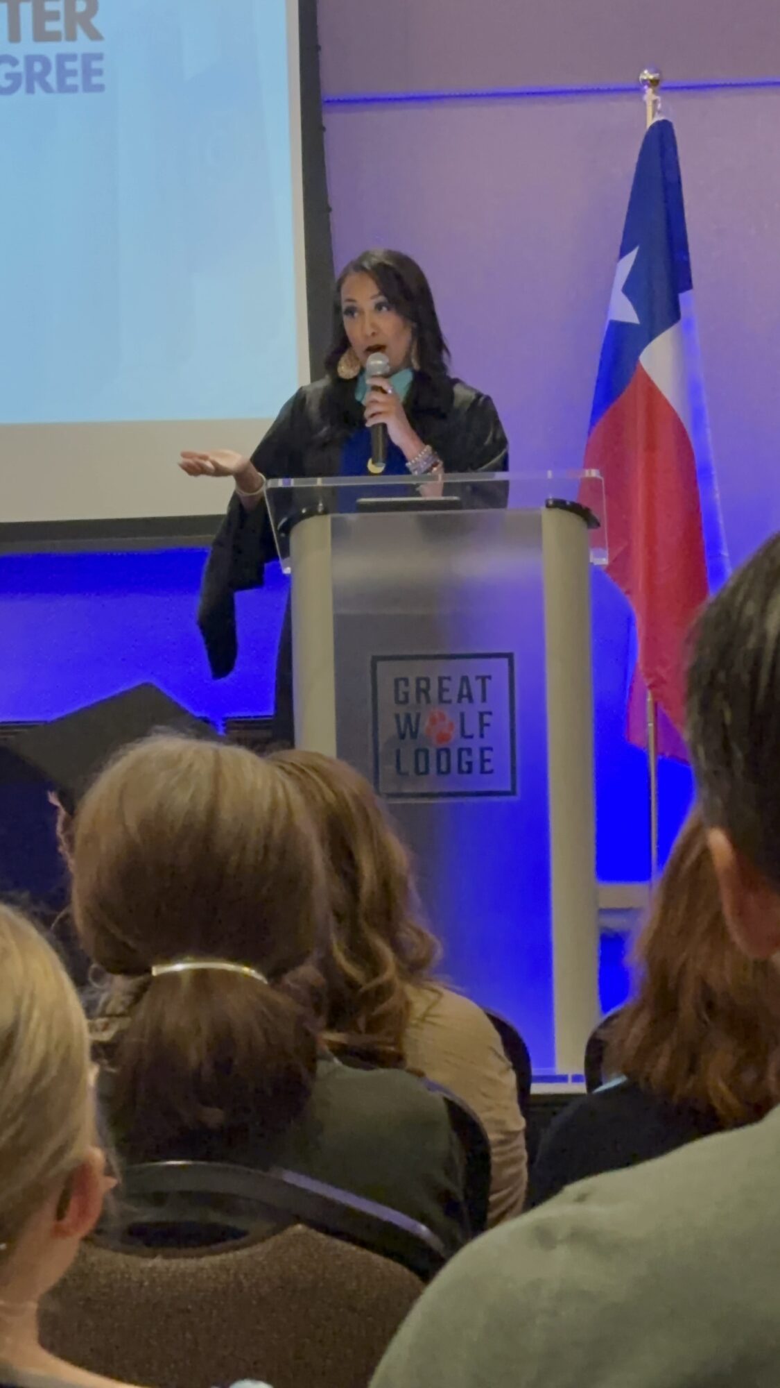 Woman speaking at a podium with a microphone, audience in foreground, Texas flag behind, presentation screen to the left.