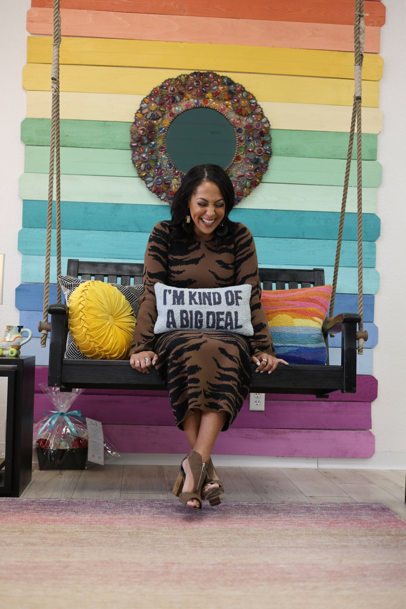 Woman standing in front of a rainbow-colored wall holding a sign, with a yellow umbrella on a bench, smiling.
