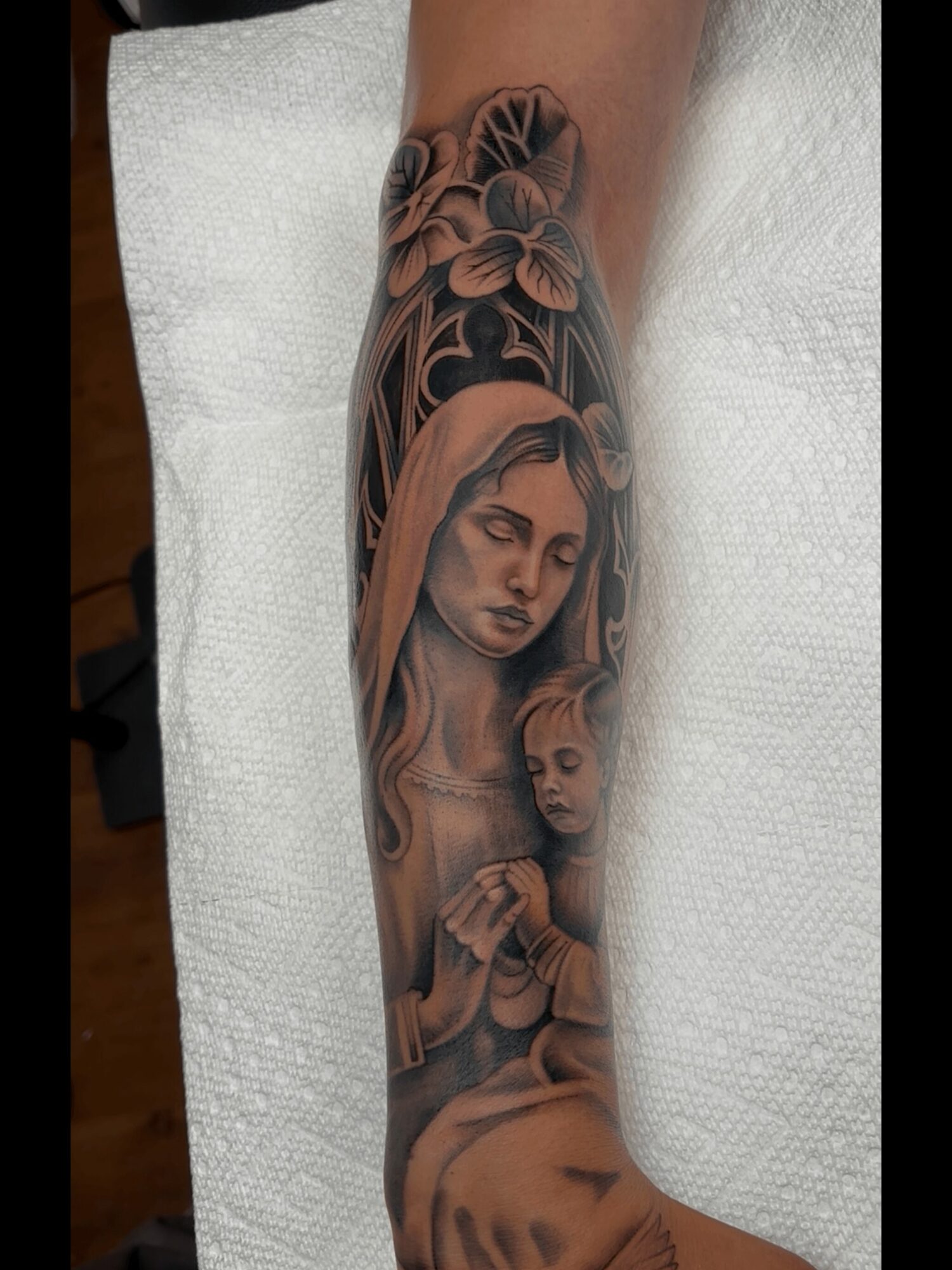 Tattoo of a woman with closed eyes and hands clasped, with a child, surrounded by flowers and decorative elements.
