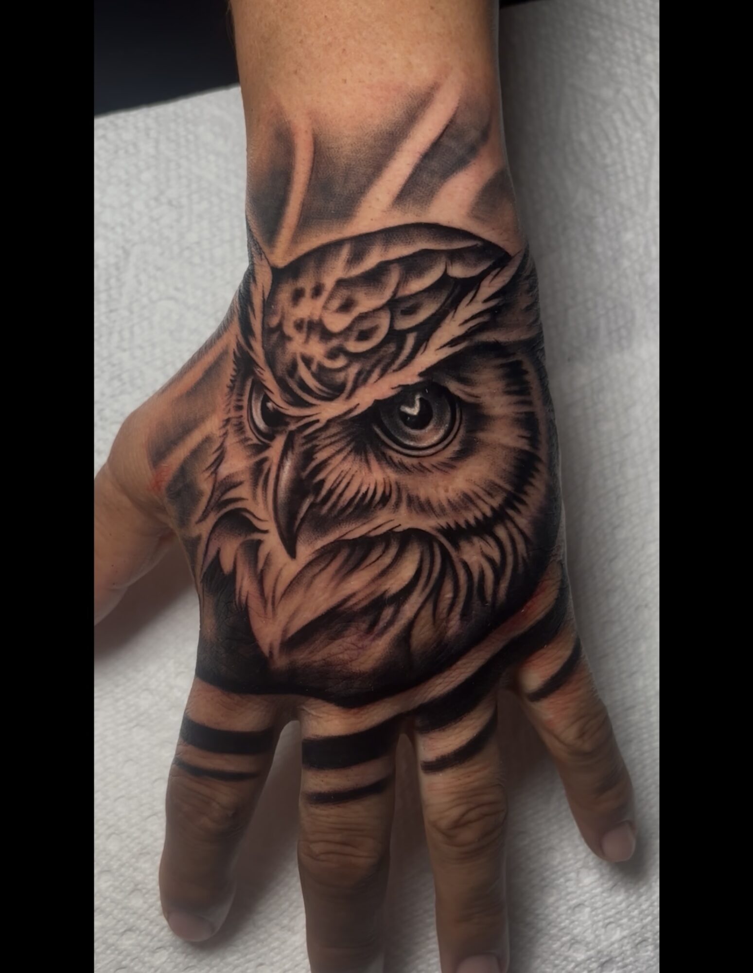 Tattoo of an owl's face on a person's hand and wrist, with detailed feathers and eyes, in black and gray ink.