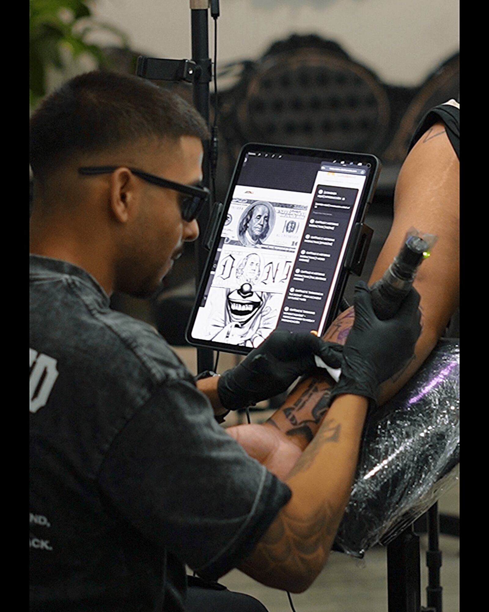 Tattoo artist in black gloves working on a client's arm with a tablet displaying design sketches.