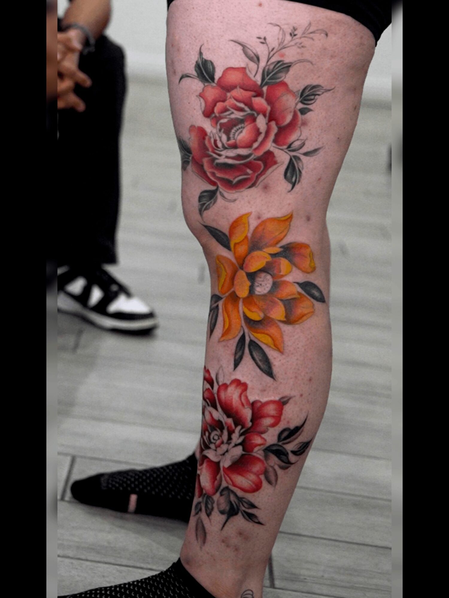 Tattooed leg with large red and orange flowers, black leaves, and detailed shading, shown on a person's lower leg.