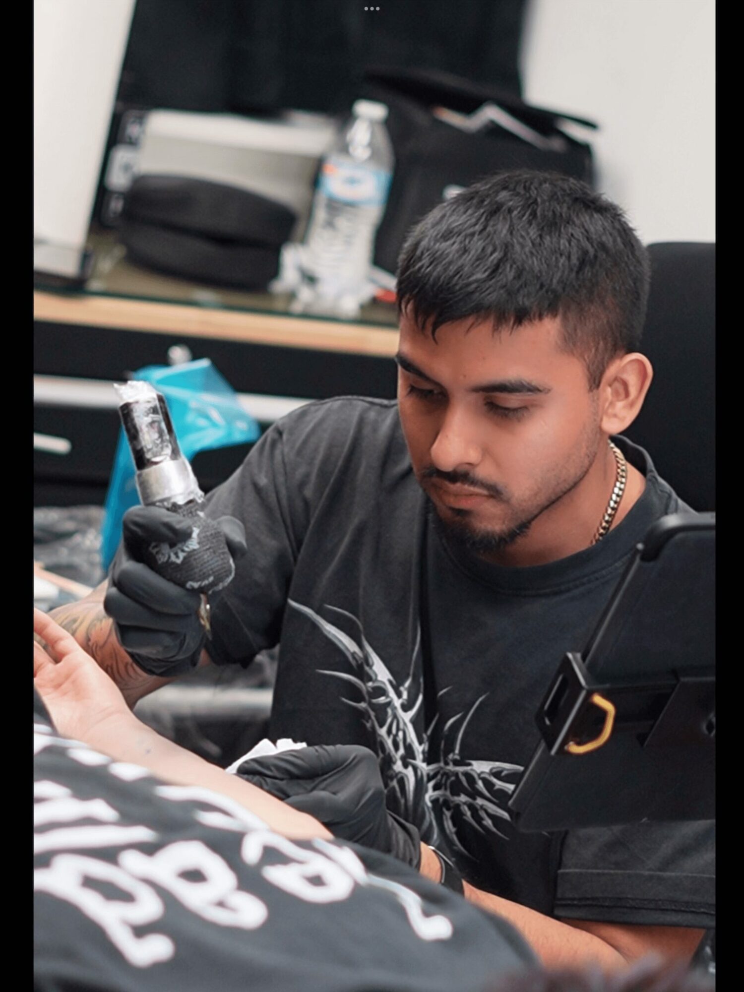 Young man with short dark hair and a beard, wearing a black shirt, holding a tattoo machine, focused on tattooing.