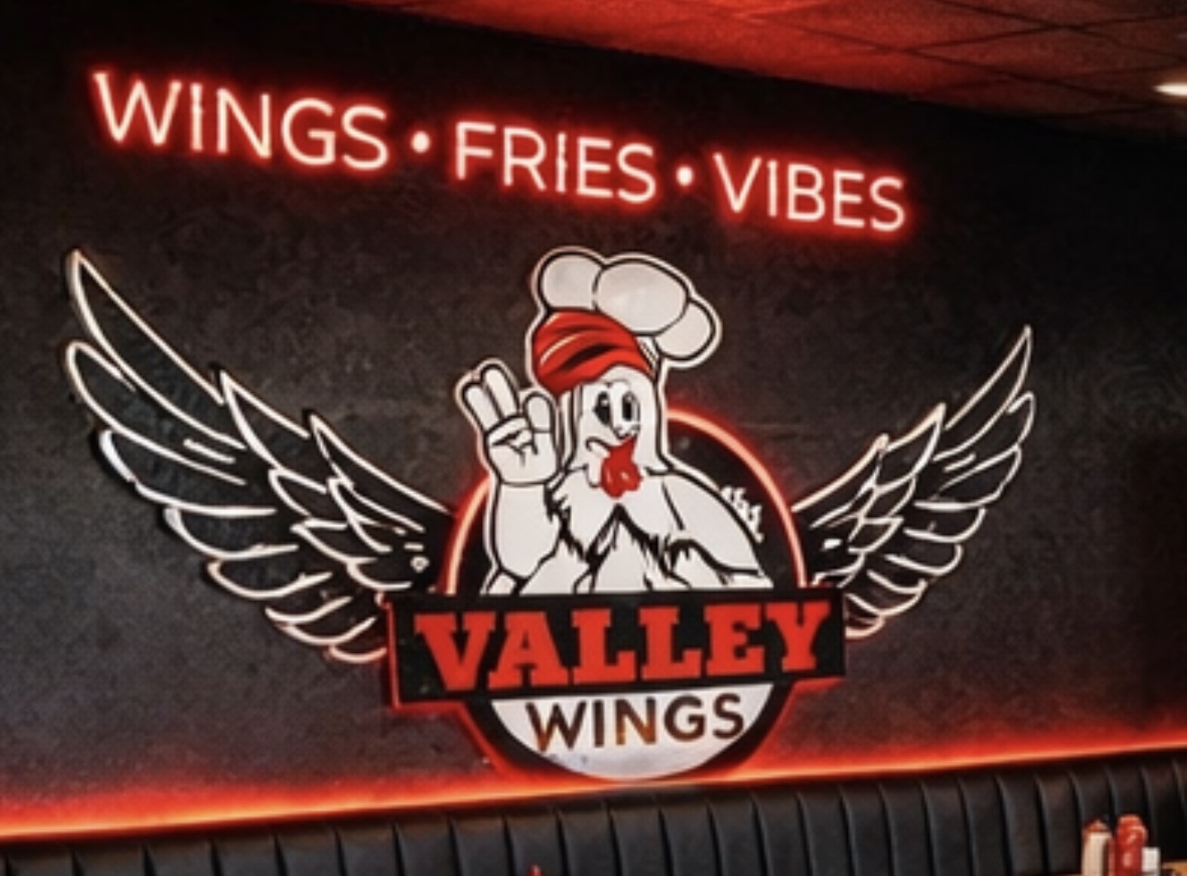 Neon sign with text 'WINGS FRIES VIBES' above a logo of a chicken with wings, wearing a chef's hat, making a peace sign, with wings spread wide.