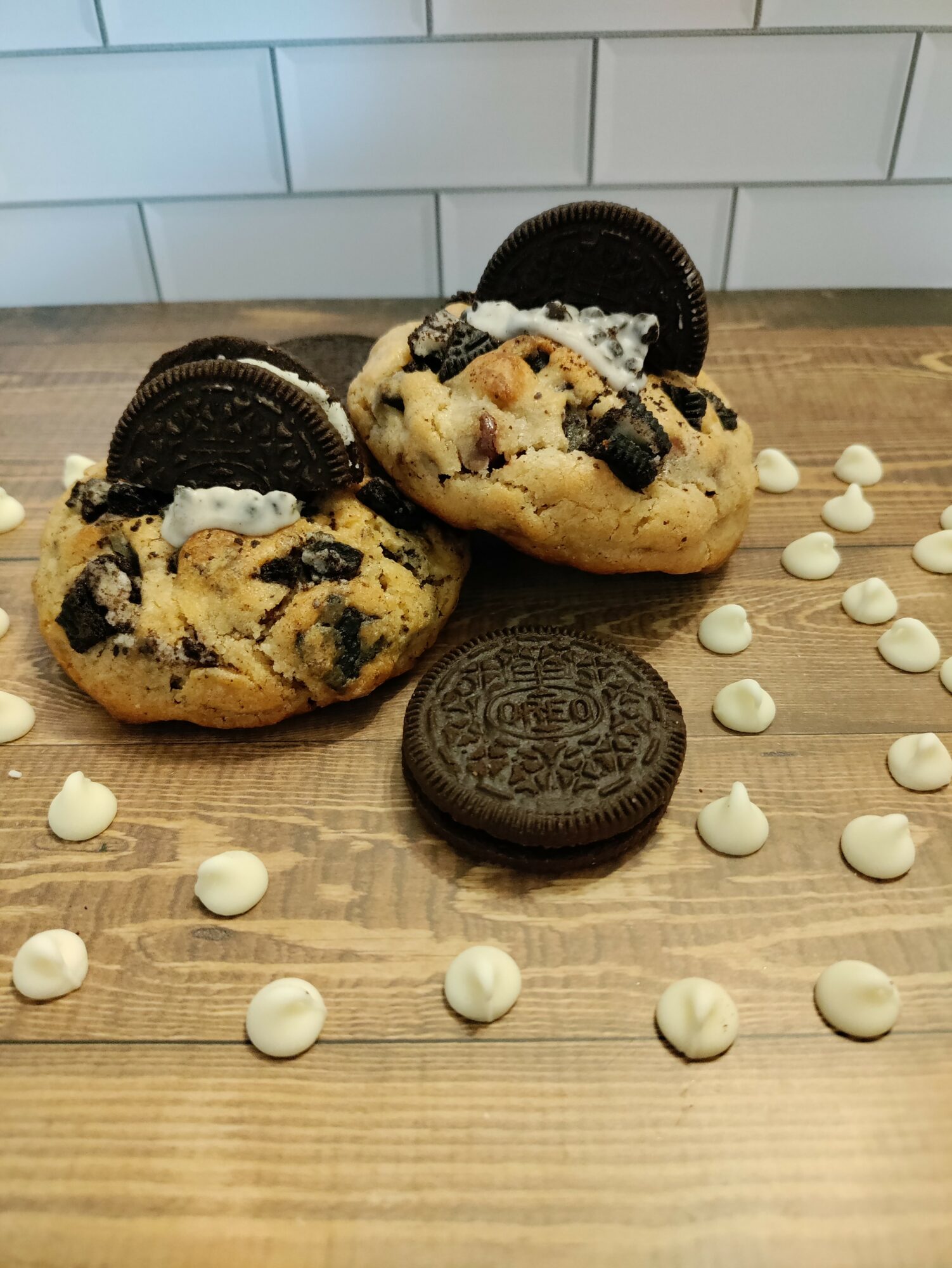 Two cookies with Oreo pieces and cream, one with an Oreo cookie on top, surrounded by scattered white chocolate chips on a wooden surface, with a white tiled wall in the background.
