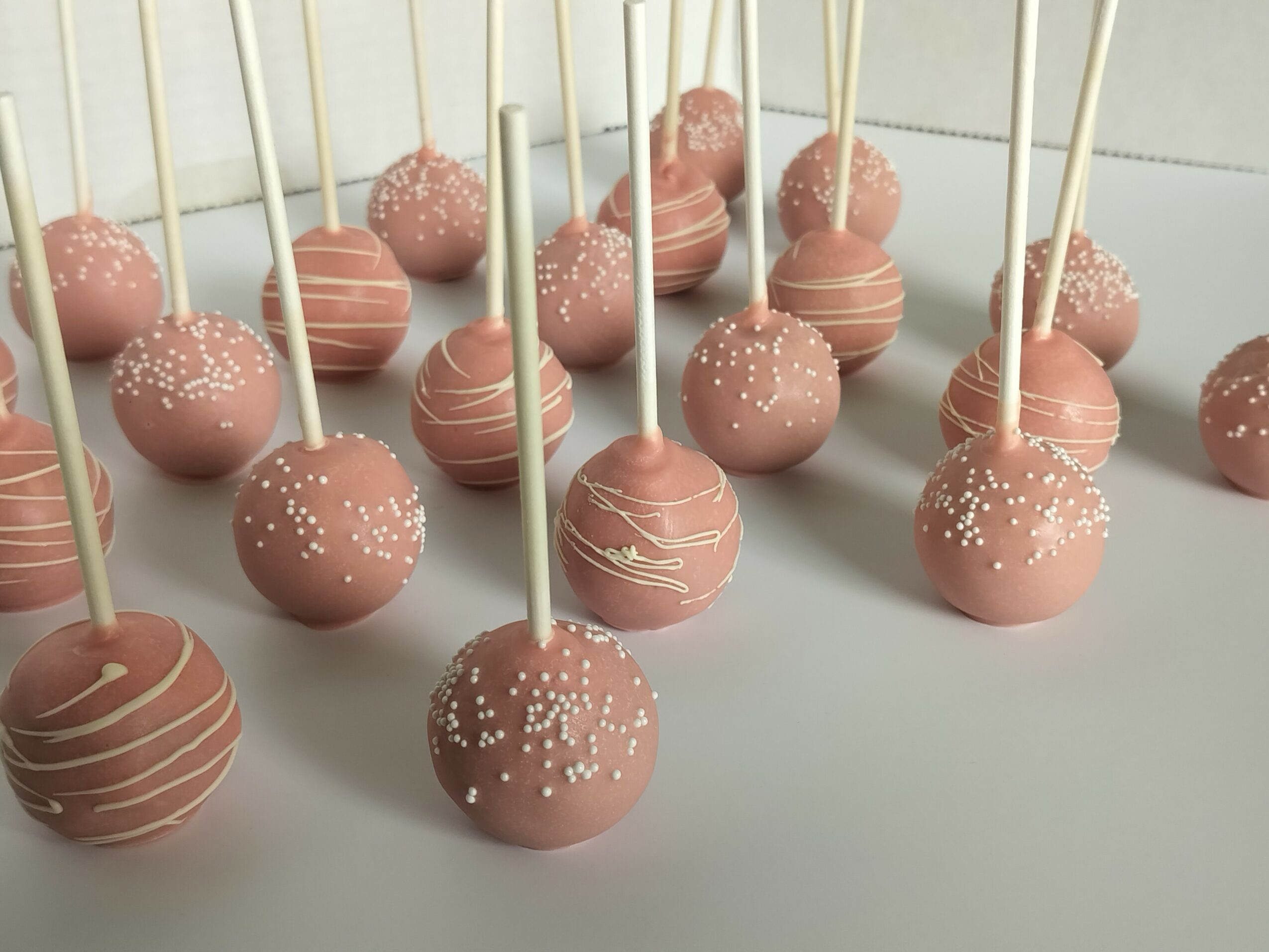 Pink cake pops with white sprinkles and drizzle, arranged on a white surface, each with a stick, in a close-up view.