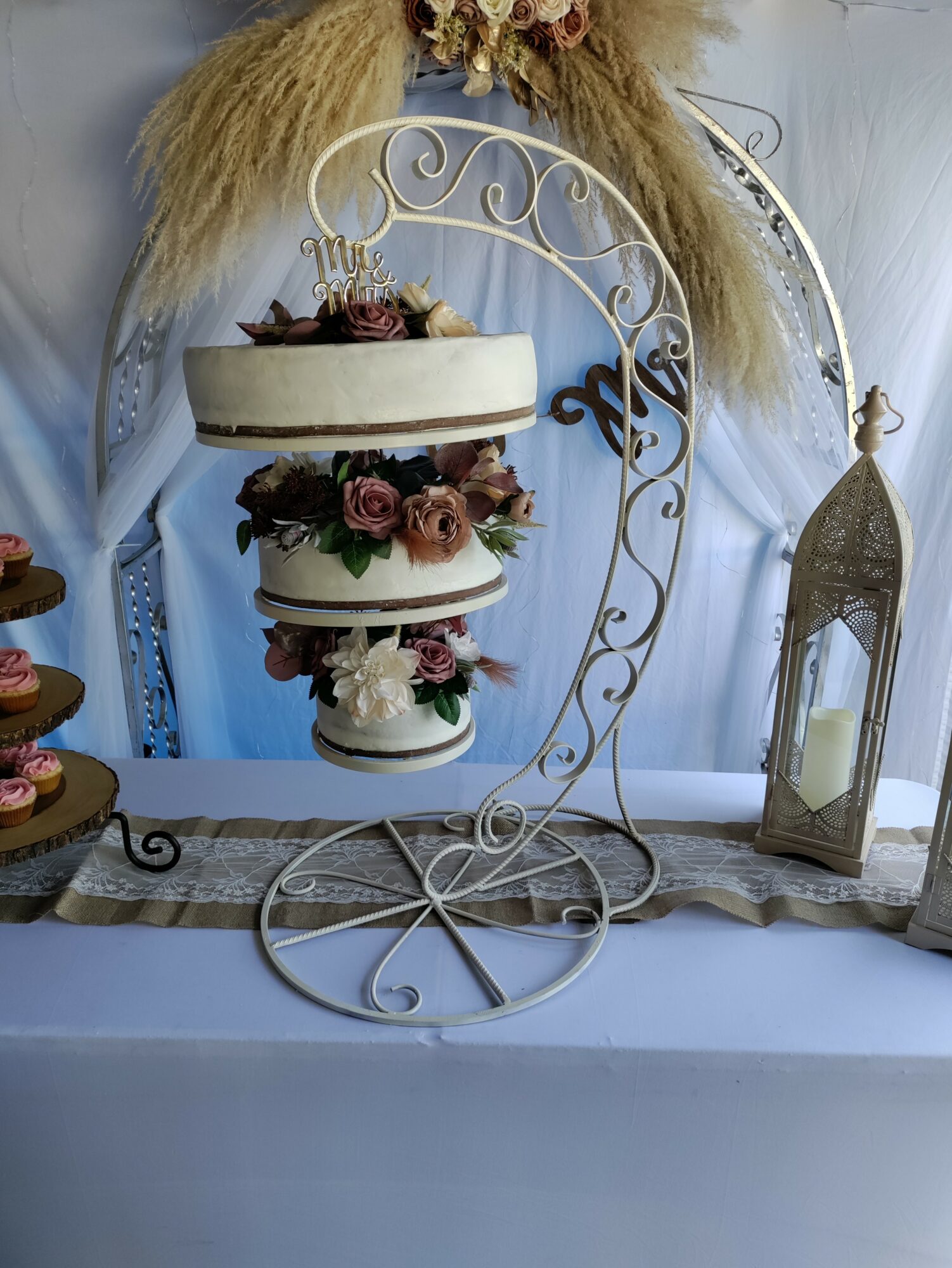 Decorative display with tiered cake stand, flowers, and a lantern on a table, with floral arrangements and wheat in the background.