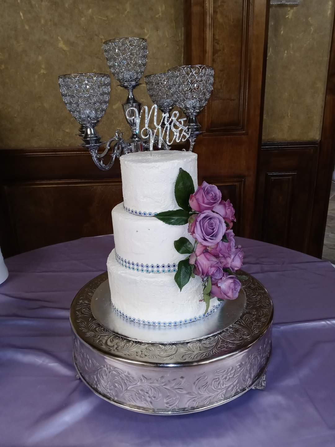 Two-tier white wedding cake with purple roses and green leaves, topped with a silver 'Mr & Mrs' cake topper, on a silver stand.