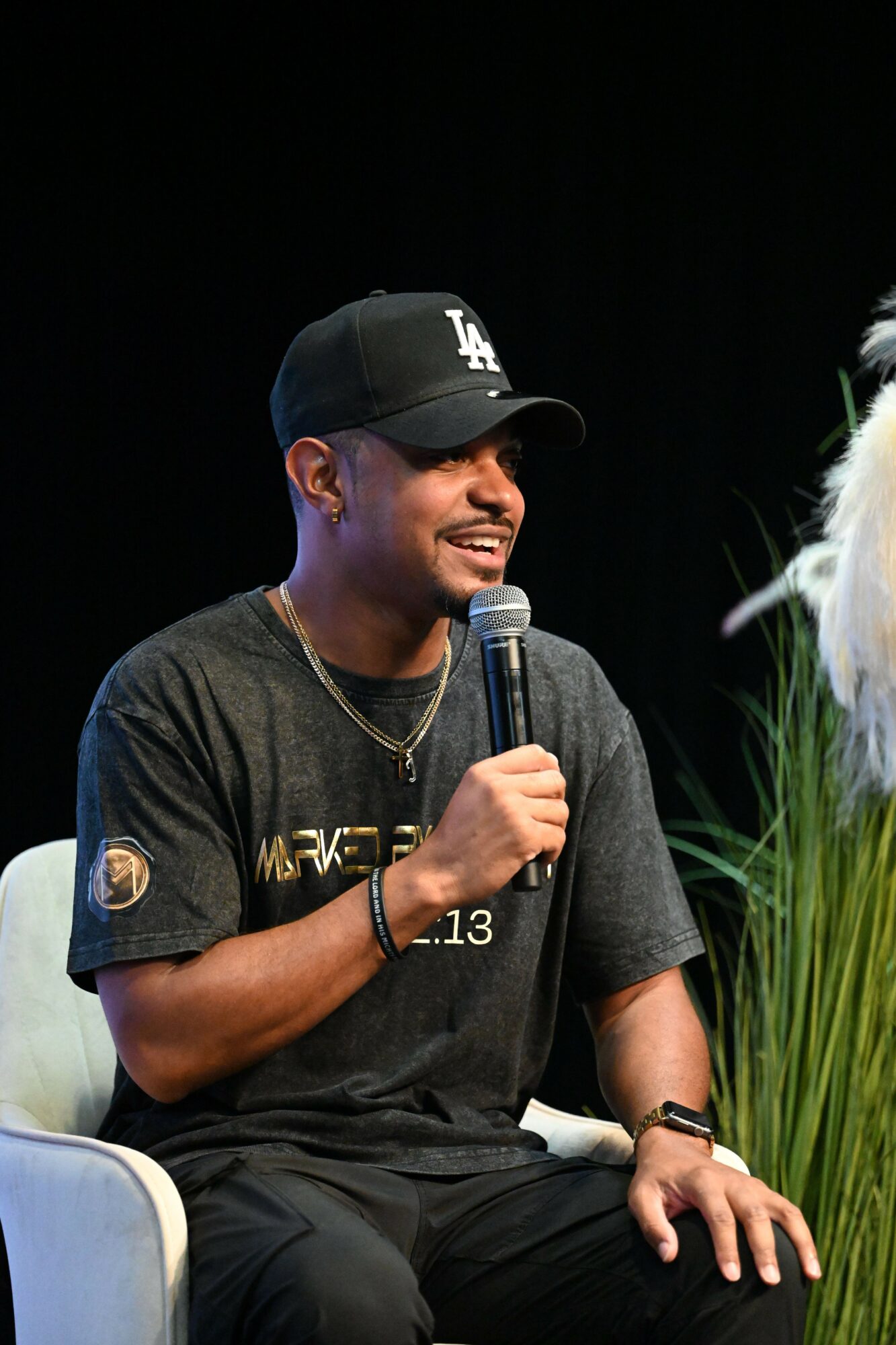 Person wearing a black cap and dark clothing, holding a microphone, smiling, with green plants in the foreground.
