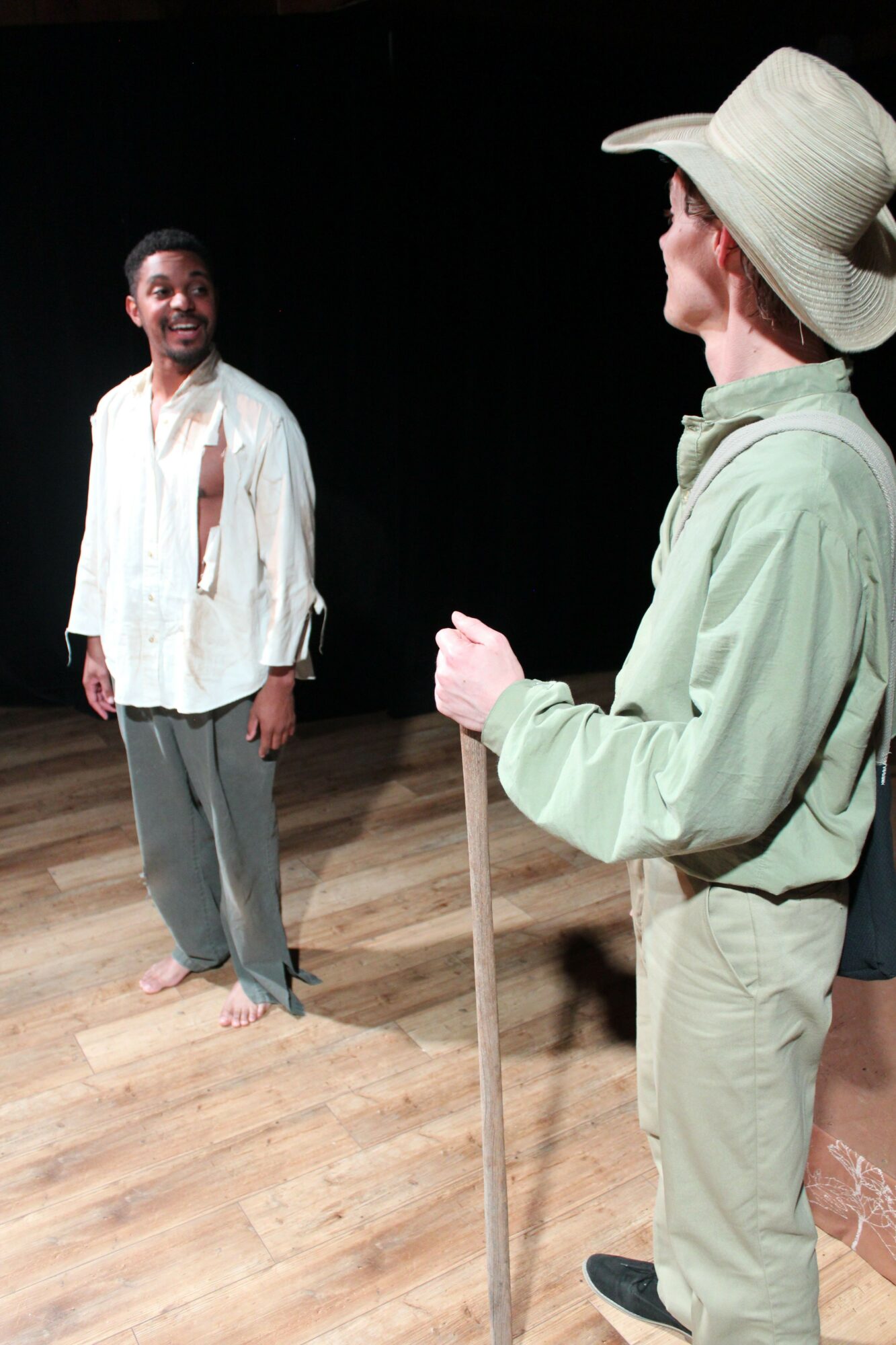 Person standing on a wooden floor, wearing a wide-brimmed hat, light green jacket, and beige pants, with a dark background.