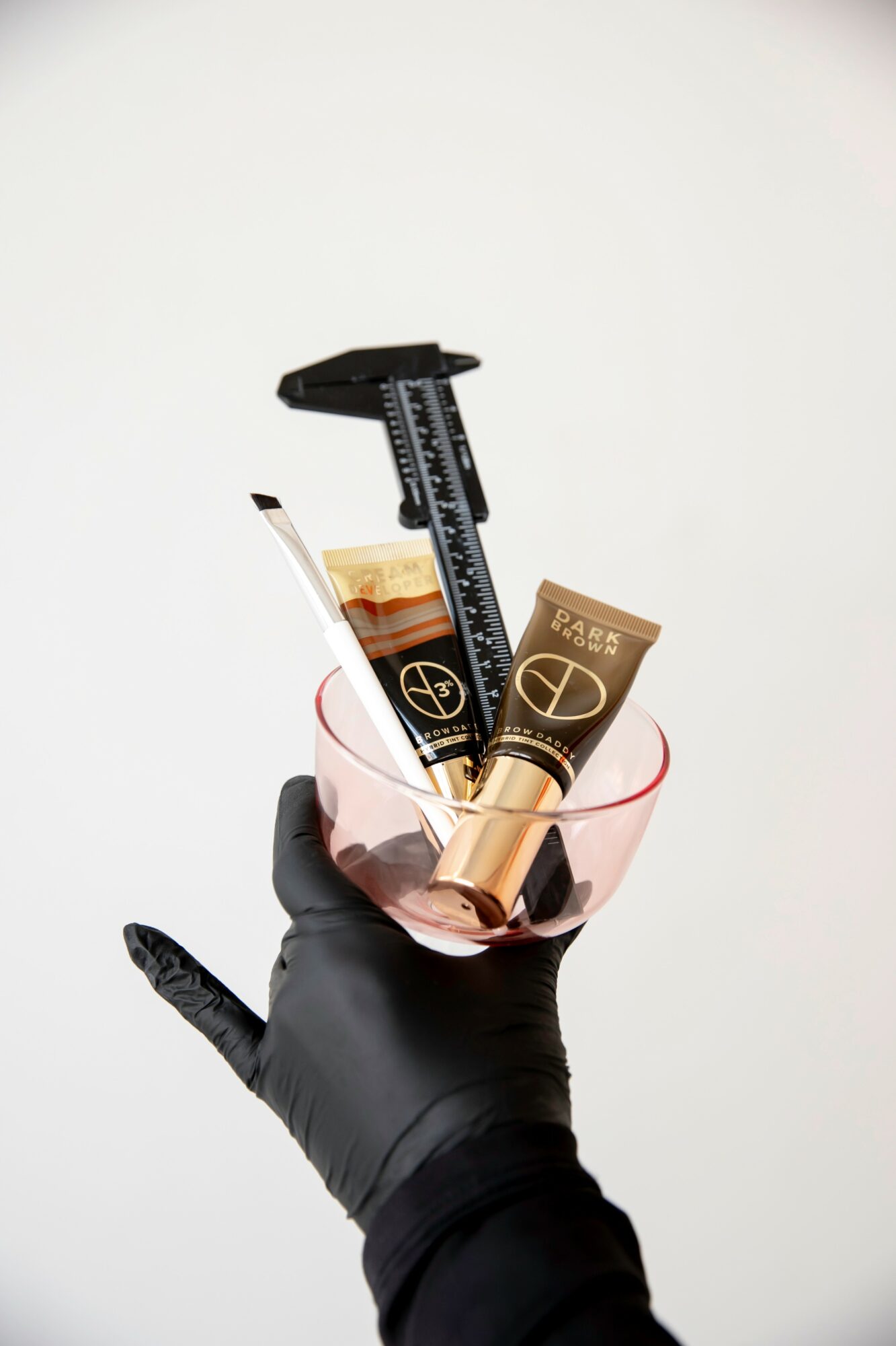 Hand wearing a black glove holding a glass with various makeup products inside, against a plain background.