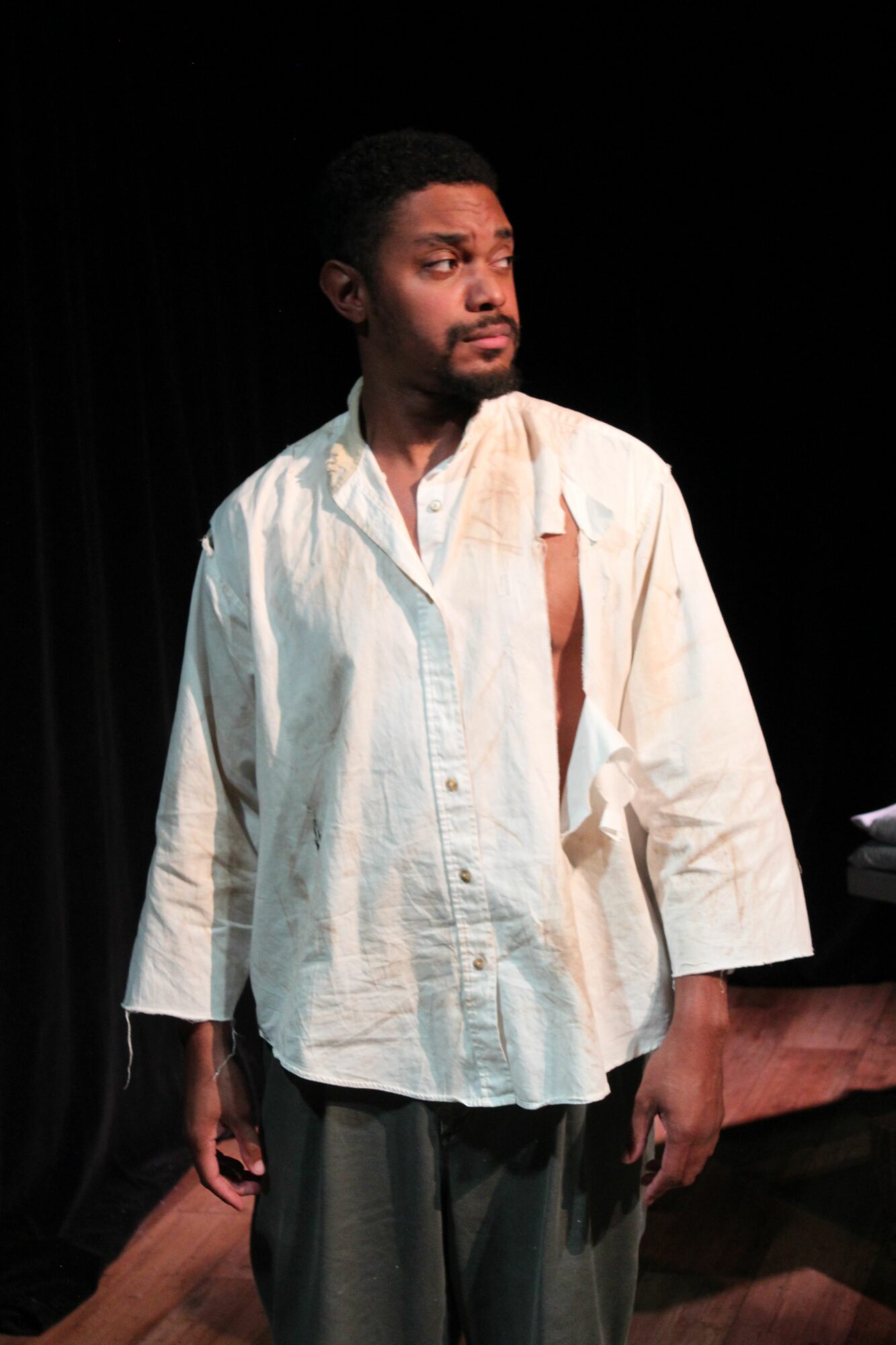 Person standing on stage wearing a white shirt and dark pants, with a dark background behind them.